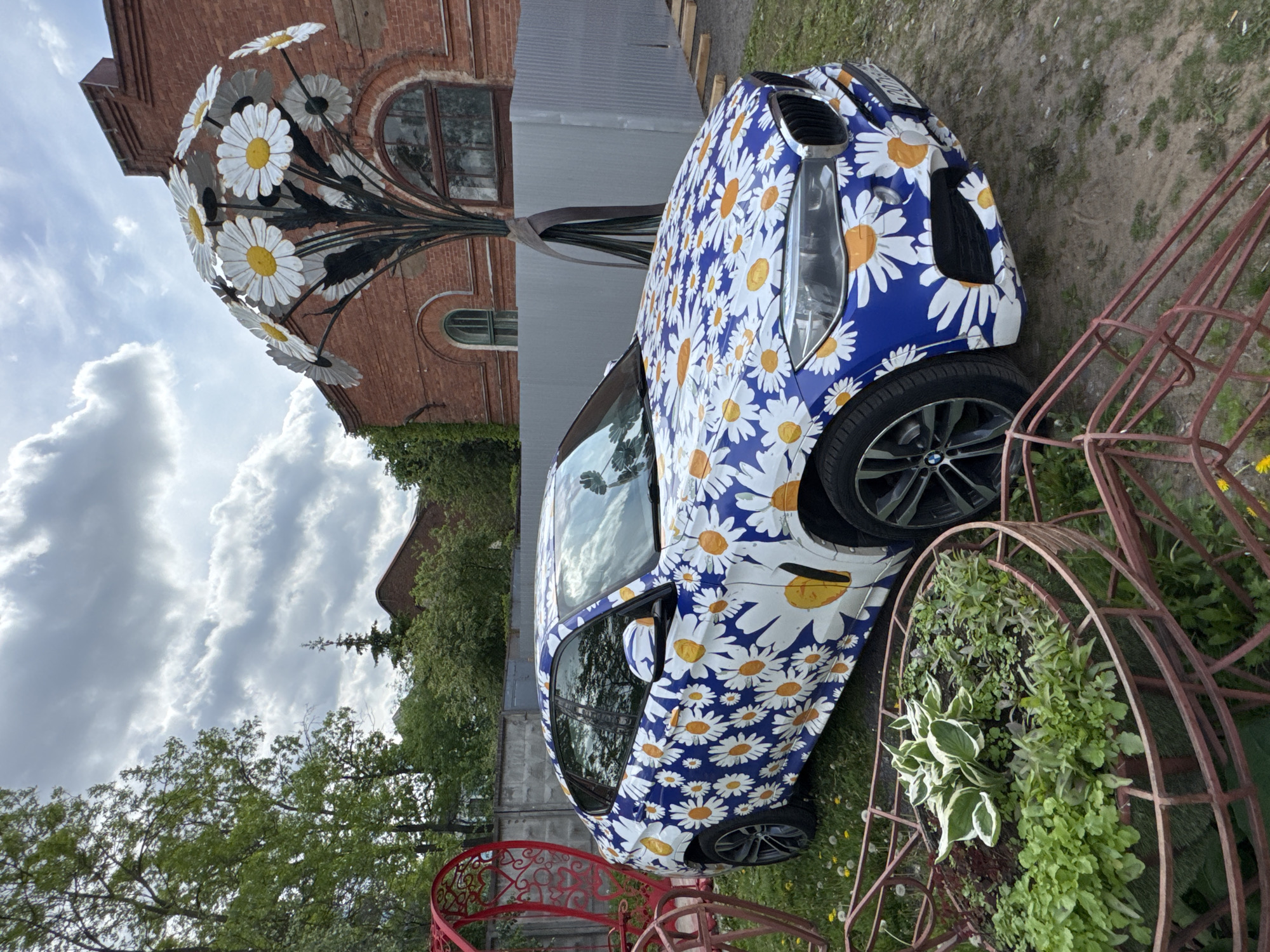 This media file presents an outdoor scene in Saint Petersburg, Russia, centered around a unique artistic display. The main subject is a compact car, possibly a BMW or Mini Cooper model, completely customized with a vibrant daisy pattern. The car's body is predominantly blue, covered with numerous white daisies featuring bright yellow or orange centers. It is parked on a patch of grass and dirt.
To the left and behind the car stands a large, intricate metal sculpture resembling an oversized bouquet of daisies. These sculpted flowers have white petals and yellow centers, supported by dark, metallic stems and leaves, suggesting a wrought-iron construction. The sculpture towers above the car, serving as a prominent artistic focal point.
In the foreground and to the right, a rusty, ornate metal structure, possibly a decorative planter or garden archway, contains various green plants and foliage. The surrounding ground is grassy with scattered dandelions. In the background, there are brick buildings, some featuring arched windows indicative of older architectural styles, alongside a simpler gray building. The sky is overcast with noticeable clouds, suggesting daytime conditions. There is no clearly legible text visible within the frame, though some indistinct white text may be present on the rear of the car. The overall setting evokes an artistic or garden exhibition space.