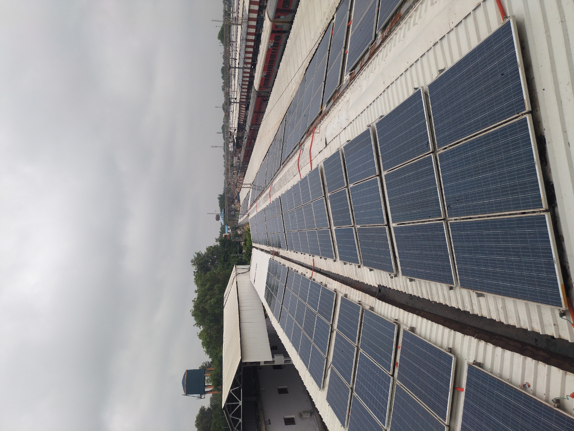 The image captures a view from an elevated vantage point, looking over the rooftop of a building covered in solar panels. The solar panels are arranged in multiple rows and are dark blue in color with silver frames. They are installed on a white, corrugated metal roof. In the background, train tracks are visible with several red and white passenger trains parked along them. Beyond the train tracks, a line of trees can be seen, and further in the distance, there are some buildings and structures, including a water tower. The sky is overcast and grey. The scene appears to be a railway station in Agra, India, likely a railway yard or maintenance area due to the presence of multiple trains and the large scale installation of solar panels on the rooftops of adjacent buildings. There are no people visible in the image. The overall impression is one of urban infrastructure development focusing on renewable energy.
