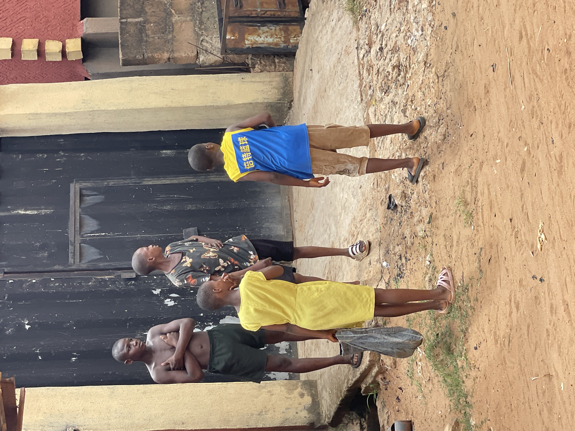 Four people are standing in an outdoor area in Nkpor, Nigeria. The person closest to the camera is shirtless and wearing dark green shorts. He has his arms crossed. The next person is wearing a yellow dress. The third is wearing a dark shirt with butterfly patterns and black shorts. The farthest person is wearing a blue shirt with yellow trim and khaki shorts. They are standing near a black wall, next to a weathered tan wall, and a patch of ground with dirt.