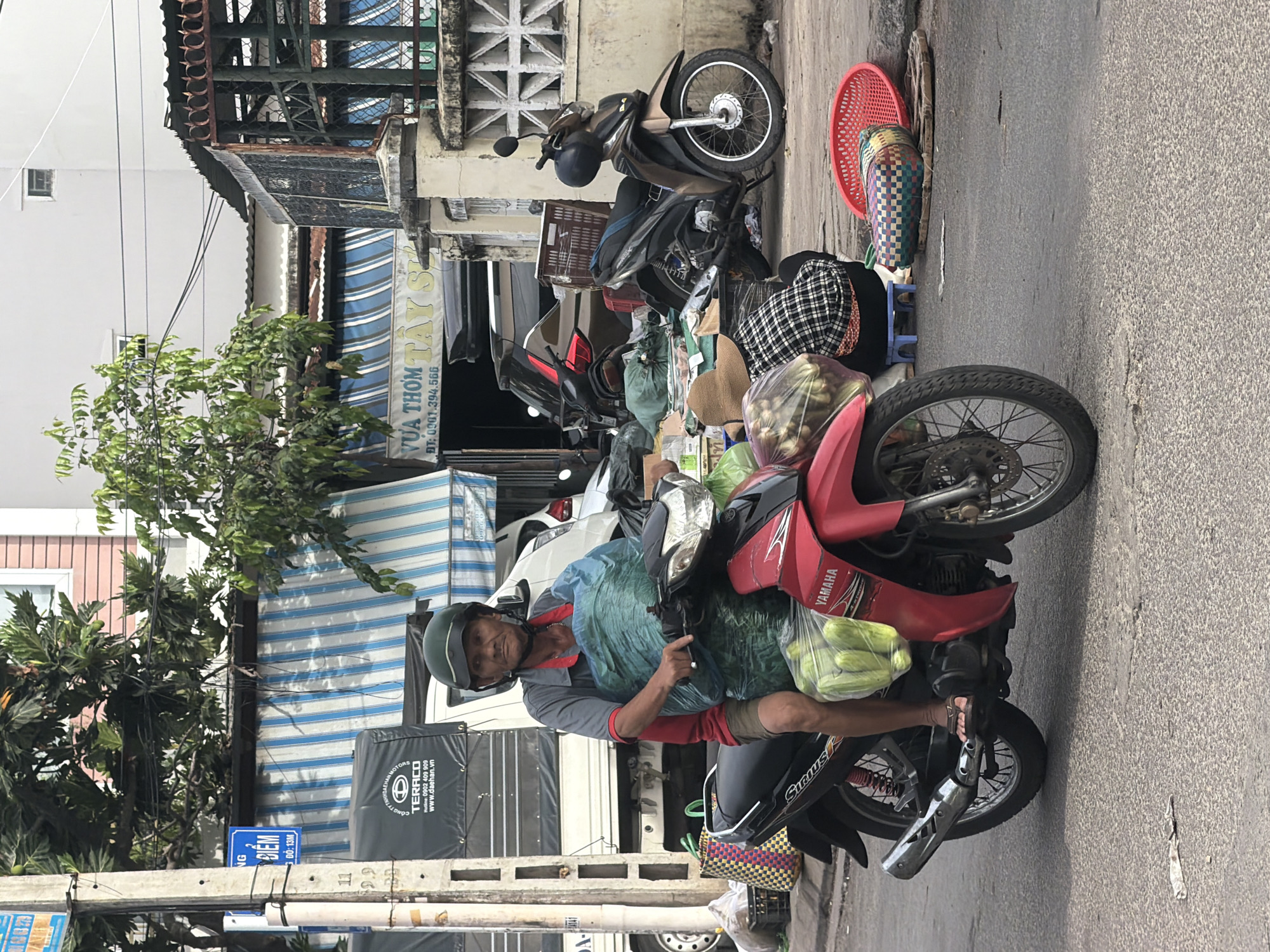 The image shows a man riding a red Yamaha motorcycle on a city street in Nha Trang, Vietnam. He is wearing a green helmet and a grey shirt with red trim. He is carrying a large blue bag of what appears to be produce on the motorcycle in front of him. He also has a plastic bag of green items hanging from the front of the motorcycle. The man is looking towards the camera. In the background, there are buildings, a truck, another motorcycle, and several woven baskets and mats on the ground. There is also a blue sign on a pole with text in Vietnamese. The street appears to be paved with asphalt. The lighting suggests it is daytime.