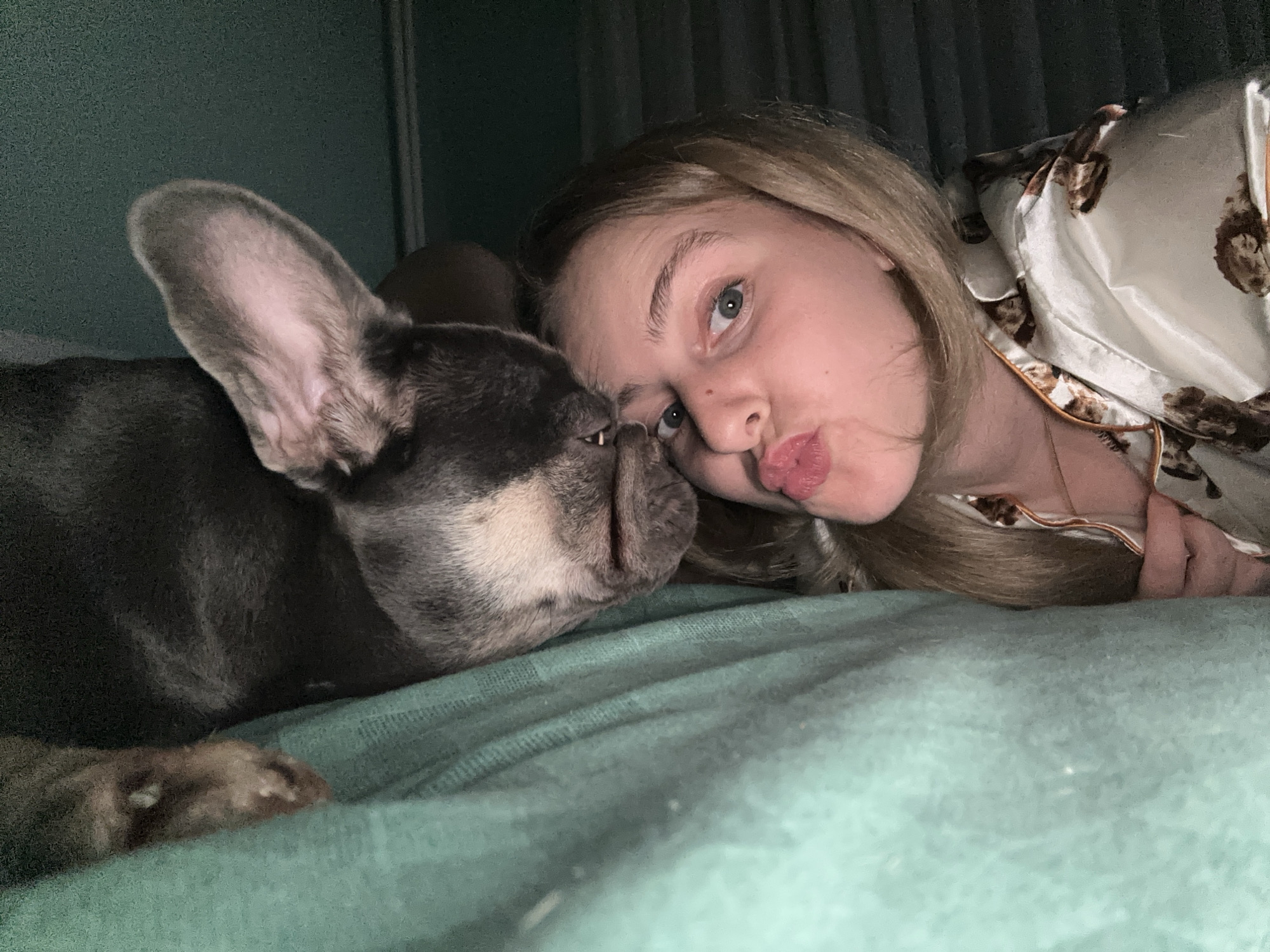 This close-up media file captures an intimate moment between a young person and their French Bulldog. The subjects are lying down on a teal-colored surface, likely a bed, with a person in the foreground and the dog snuggled close to them.
The young person, with blonde hair, is looking towards the camera with a playful expression, puckering their lips for a kiss. They are wearing a light-colored pajama top with a subtle print. The French Bulldog is dark gray with a lighter muzzle and large, prominent ears. Its head is resting against the person's face, creating a sense of warmth and affection. The dog's paw is visible in the lower left corner, resting on the teal surface.
The lighting suggests it might be nighttime or a dimly lit room, creating a cozy and private atmosphere. The background is dark and out of focus, with hints of vertical lines, possibly suggesting curtains or a headboard. There are no visible text elements in the image. The overall mood is one of tenderness and companionship.