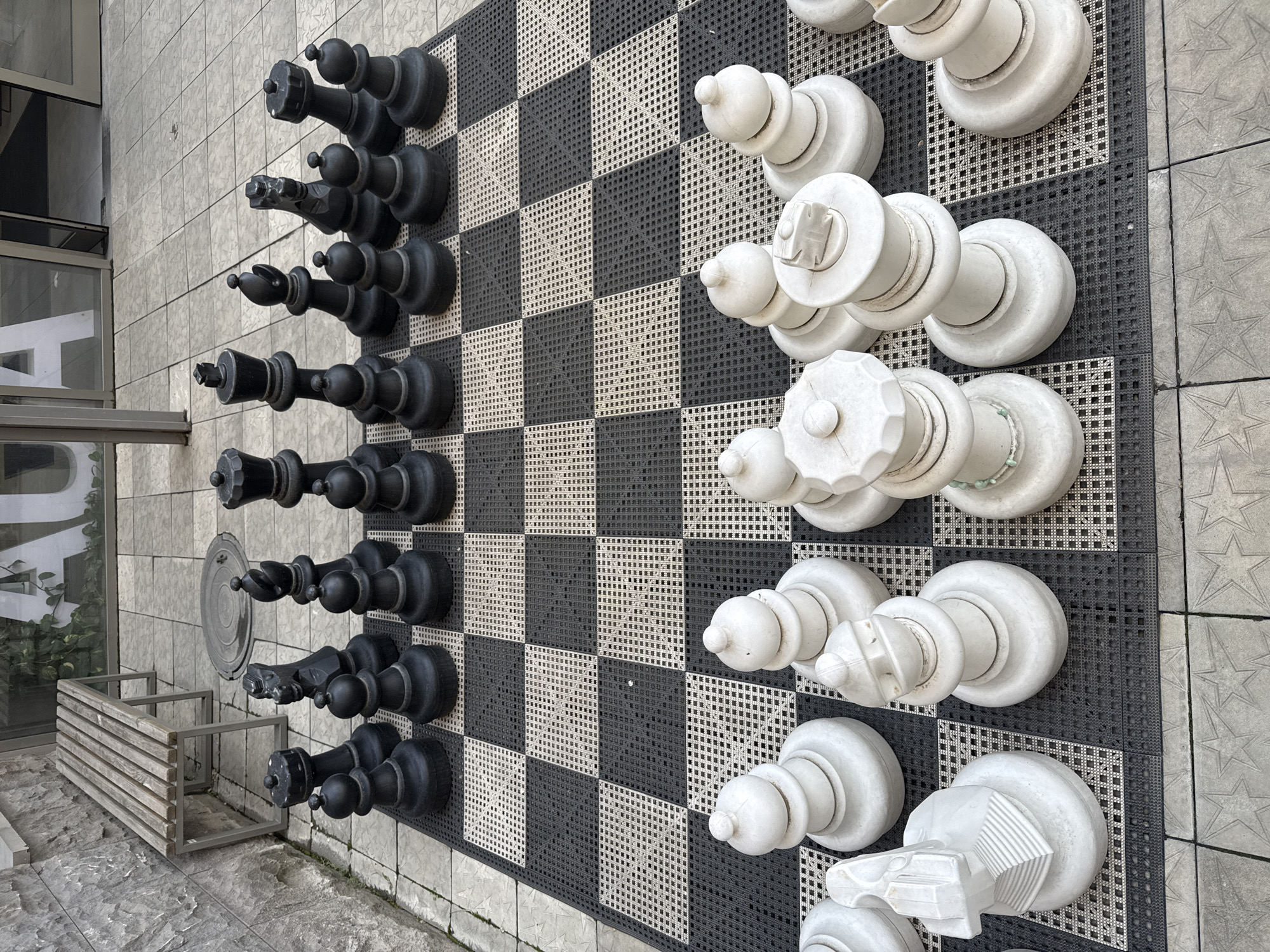 A high-angle shot depicts a large chess set with oversized pieces, positioned outdoors. The black and white checkerboard playing surface is arranged on tiled flooring. Black chess pieces are aligned on one side, while white pieces are on the opposite side. A wooden bench is located near the chess board. The scene is located in Yerevan, Armenia.