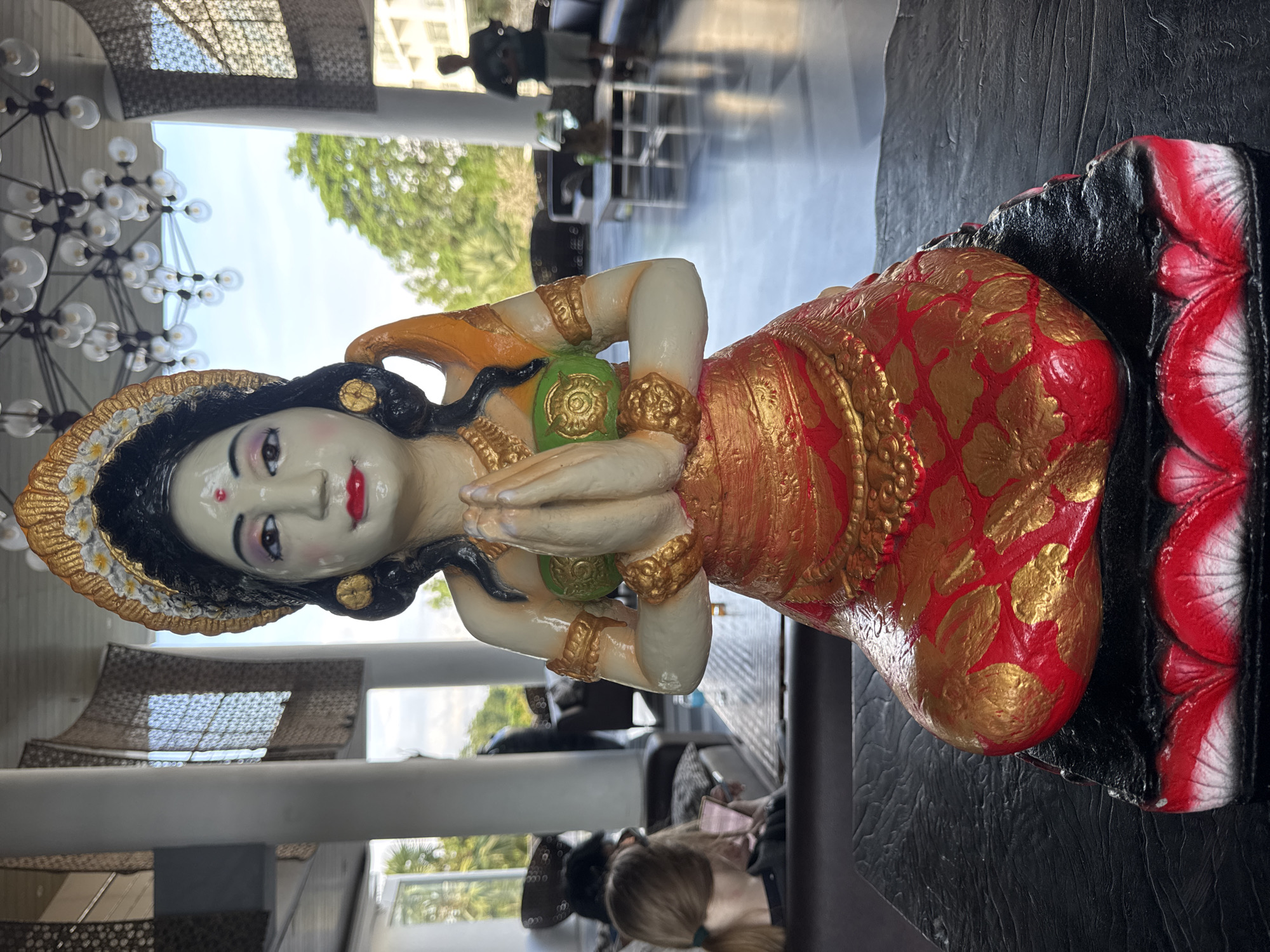 The image depicts a statue of a woman in traditional attire, adorned in red and gold, kneeling with her hands clasped in prayer. She has dark hair and fair skin, with painted facial features including red lips and a small red dot on her forehead.
The statue rests on a decorative base with red and black accents, positioned on a dark, possibly wooden, table. The background suggests an indoor space, potentially a lobby or lounge area. There are individuals seated in the background, and architectural elements like pillars and modern light fixtures are visible. The setting is likely in Pecatu, Indonesia, as that was stated with the prompt.