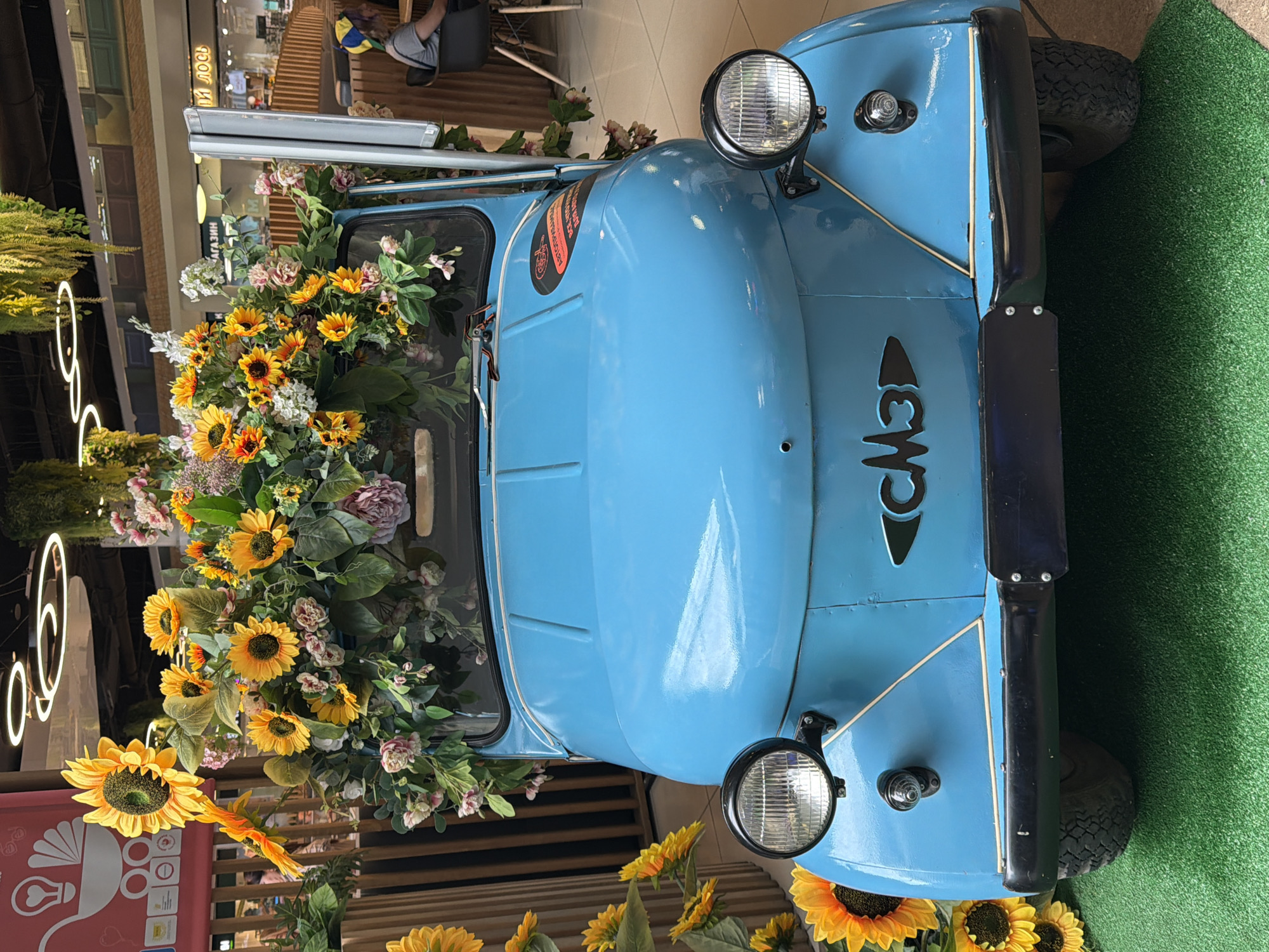 The image shows the front of a bright blue, vintage-style car that has been decorated with an abundance of artificial sunflowers and other floral arrangements. The car has a round, black bumper and two prominent headlights. A black emblem with the Cyrillic letters "СМЗ" is visible on the hood. The car is parked on a patch of artificial green turf. In the background, there are glimpses of a commercial interior, including wooden architectural elements, hanging plants, and bright ceiling lights. People are visible in the background, including one person sitting at a table and another walking by. The overall scene appears to be a decorative display, possibly in a shopping mall or public space. There is no visual information in the image that suggests a specific location in Vologda, Russia.