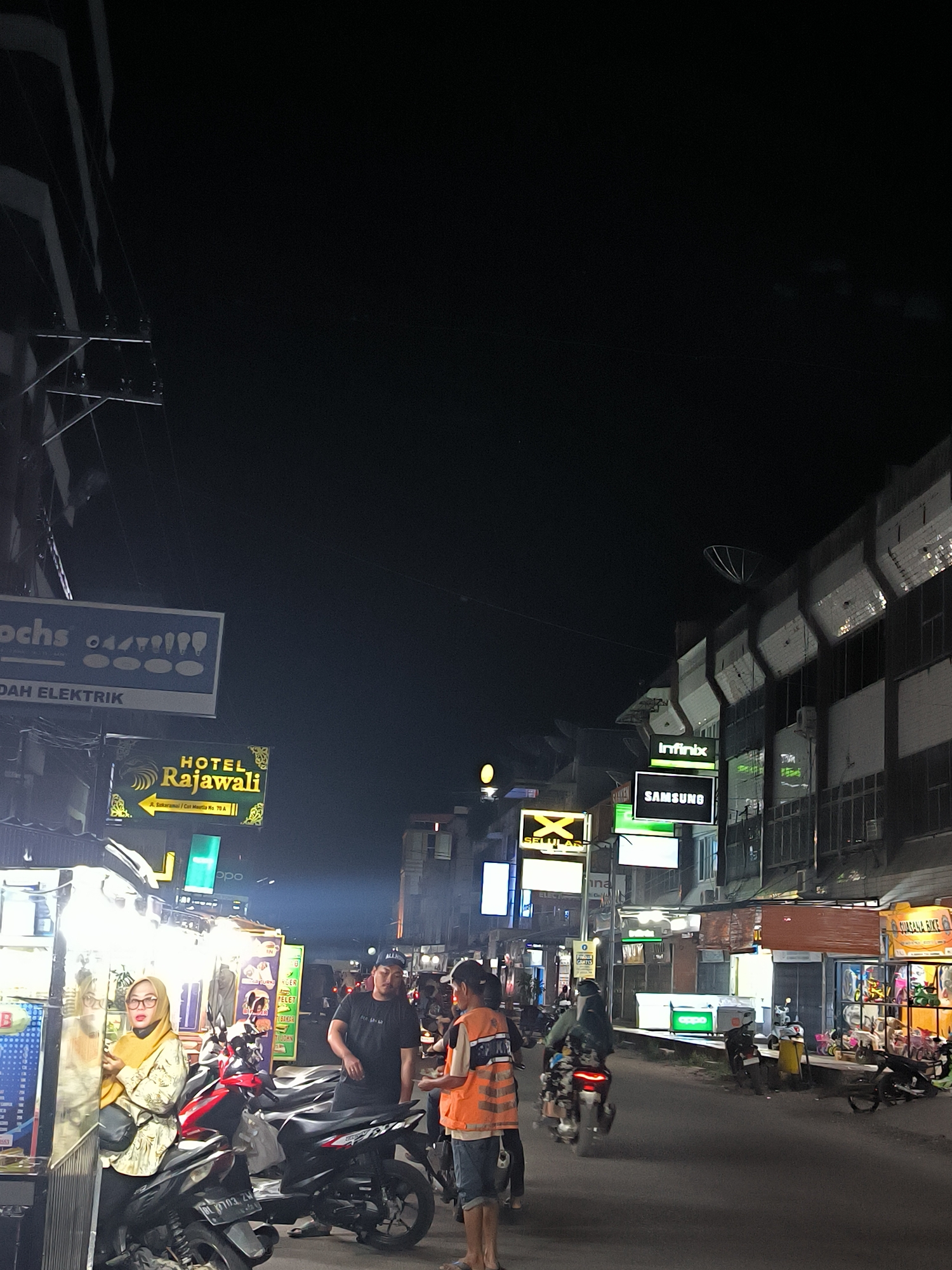 The image depicts a street scene at night in Lhokseumawe, Indonesia. The street is lined with various shops and businesses, many of which have illuminated signs. In the foreground, a woman in a yellow headscarf and patterned clothing is standing next to a motorcycle at a food stall. To her right, a man wearing a black t-shirt and a cap is standing, and further to the right, another person in an orange vest and shorts is interacting with someone off-camera. Several motorcycles are parked or being ridden on the street. Signs visible include "Ochs" and "DAH ELEKTRIK", "HOTEL Rajawali", "Infinix", "SAMSUNG", and "GUAYANG BIKE". The architecture appears to be typical of urban Indonesia, with multiple stories and commercial storefronts. The overall atmosphere is that of a busy, active night market or commercial street.