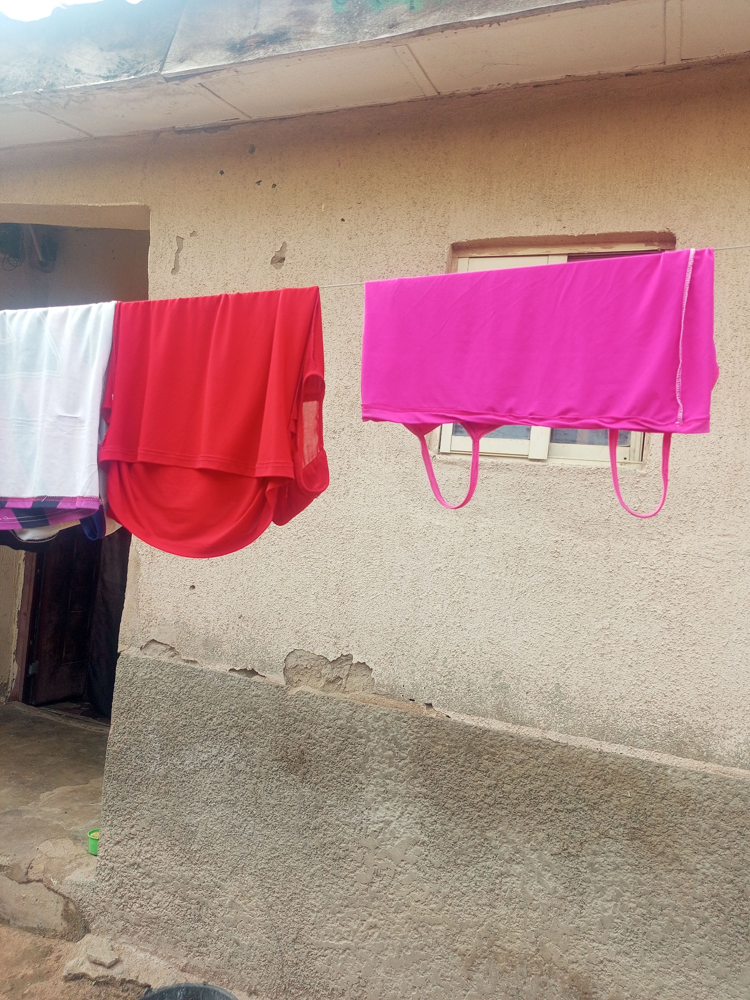The image captures an outdoor scene in Jalingo, Nigeria, where laundry is hanging on a clothesline stretched across the front of a building. The building's exterior wall is a textured, light-colored stucco, showing signs of wear and tear with peeling sections. A small, rectangular window with light-colored blinds is visible in the wall. Three garments are drying on the line. On the left, a white garment with faint patterns is partially visible. Next to it is a bright red garment, draped in folds. To the right, a vibrant pink garment, possibly a tank top or dress, is hanging with two loops visible at the bottom. The clothesline appears to be suspended against the building's facade, with the roofline of the building visible at the top of the frame. The ground in the foreground consists of uneven, earth-toned surfaces, with some concrete elements and a small green bucket visible near the building's base.