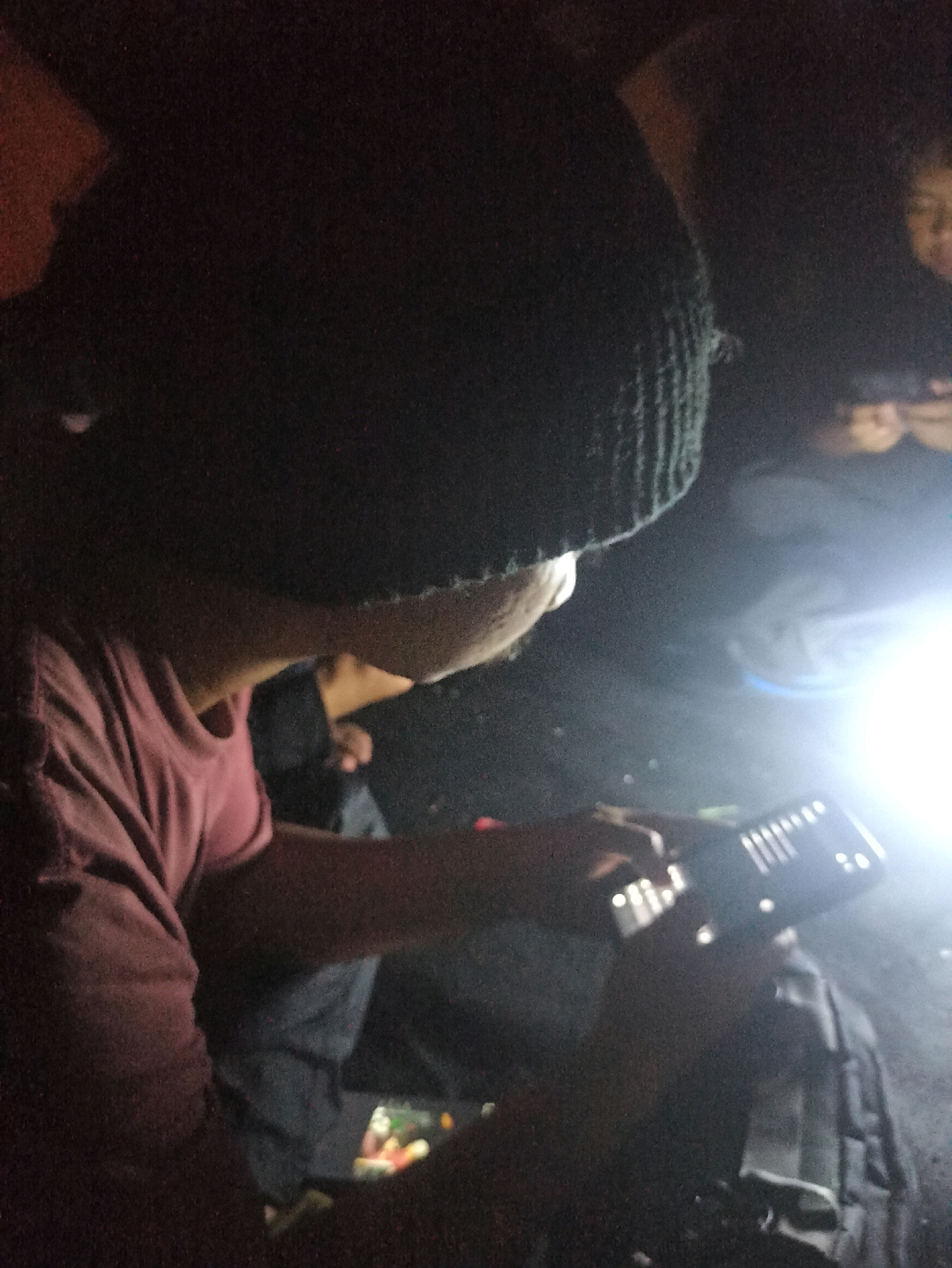 In low light conditions, possibly at night, an individual wearing a dark beanie and a maroon t-shirt is focused on a tablet computer held in their hands. The tablet emits a bright light, illuminating the surrounding area and partially obscuring other individuals and details in the background. The scene appears to be outdoors, with what might be a backpack visible to the right of the individual using the tablet. The presence of a bright light source suggests a potentially portable or handheld device being used for illumination or as a primary focus of activity in a dark environment. The overall atmosphere is dimly lit and intimate, with the focus primarily on the person and the illuminated tablet. Without further context, specific details about the location within Cileunyi, Indonesia, cannot be determined.
