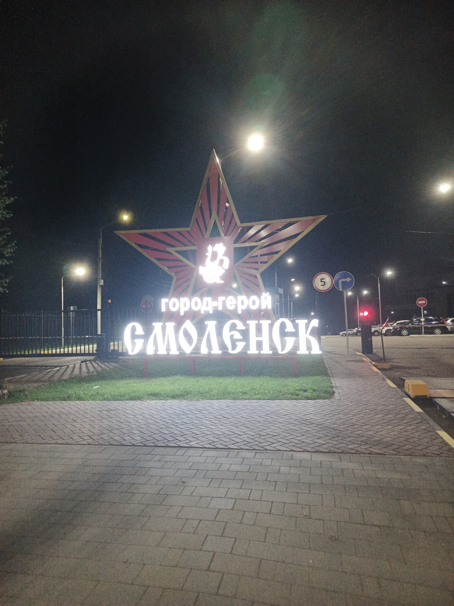 A nighttime, eye-level, medium shot captures a large, illuminated star monument in the city of Smolensk, Russia. The monument, which is red and white, features the words "Город-герой" (Hero City) and "СМОЛЕНСК" (Smolensk) in large, glowing white letters. The star is positioned against a dark night sky, with streetlights visible in the background. To the left of the monument, there is a black metal fence and some green grass. The foreground is a paved walkway made of brick-like stones. Further in the background, a road with cars and traffic lights is visible. The overall scene suggests an urban setting at night, with the star monument serving as a prominent landmark.
