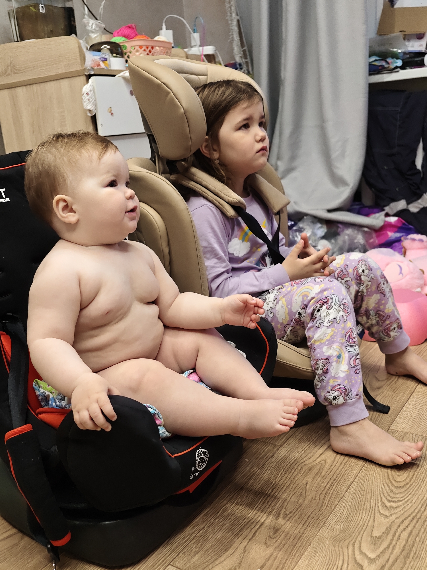 The image shows two young children sitting in child safety seats in what appears to be an indoor setting in a city of Chaikovsky, Russia. The younger child, a baby, is seated in a black car seat with a red and orange trim; the baby is unclothed from the waist up. The older child, a girl, is wearing lavender pajamas with unicorn graphics and a matching long-sleeved shirt. The girl is seated in a beige car seat with her hands clasped together. The background shows miscellaneous household objects, including a wooden dresser, baskets, and plastic bags, suggesting a lived-in environment. The floor is wooden.