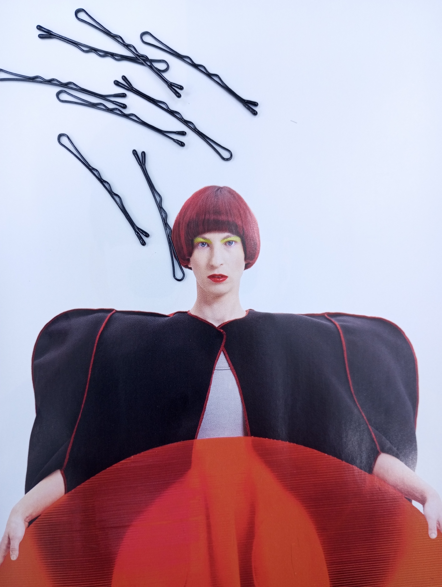 A medium shot features a person against a light blue/white background. The person has red hair in a short bob style and is wearing yellow eyeshadow. They are wearing a dark, wide-shouldered jacket with red trim and a glimpse of a light-colored shirt beneath. A translucent orange/red object or material covers the lower part of their body. Above the person's head are several black bobby pins scattered in a descending diagonal pattern. The image has a collage or cut-and-paste aesthetic. There is no visible indication of Izhevsk, Russia, or any specific location.