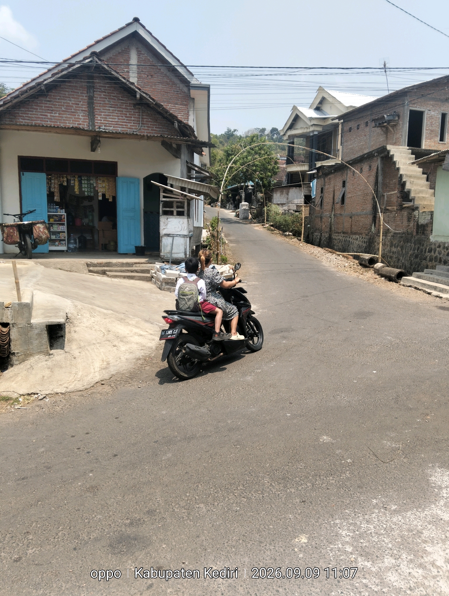 A two-person motorcycle is traveling on a paved road in Kanyoran, Indonesia. The rider, a woman with blonde hair, wears a patterned dress. The passenger, a young boy with a backpack, is seated behind her. The motorcycle is black with a red license plate that reads "AG 5385 CJ". The road is lined with brick buildings and a store with blue doors. The sky is clear and blue, indicating a sunny day. A timestamp in the lower right corner reads "oppo | Kabupaten Kediri | 2026.09.09 11:07".