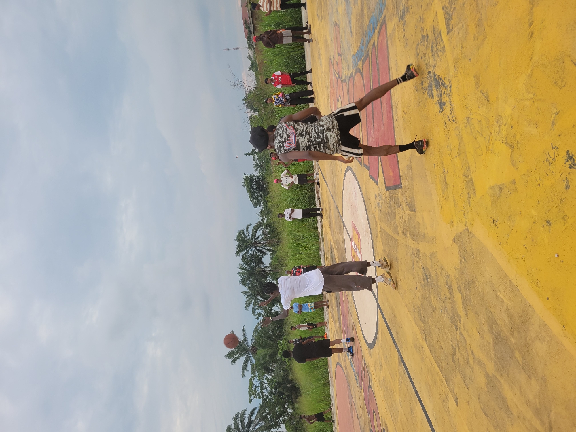 In Egbagu, Nigeria, a group of young men are playing basketball on an outdoor court. The court's surface is painted yellow with red and white markings. The sky is overcast with clouds. In the background, tall green grass and palm trees line the perimeter of the court. The men are dressed in casual athletic wear, with some wearing t-shirts and shorts, while others are in tank tops and basketball shorts. One man in the foreground is in the process of shooting a basketball. Another man, facing away from the camera, is positioned on defense. Several other men are scattered around the court, observing the game or preparing to participate. The scene captures a moment of active recreation in an outdoor urban setting.