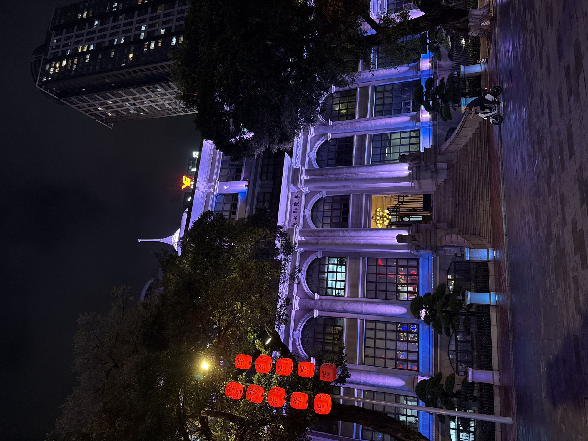 At night in Yuexiu District, China, a three-story building with rounded windows and illuminated by blue and purple lights stands alongside a taller building. A tree with red lanterns hangs in front of the building. A moped and a person are on the sidewalk next to the building.