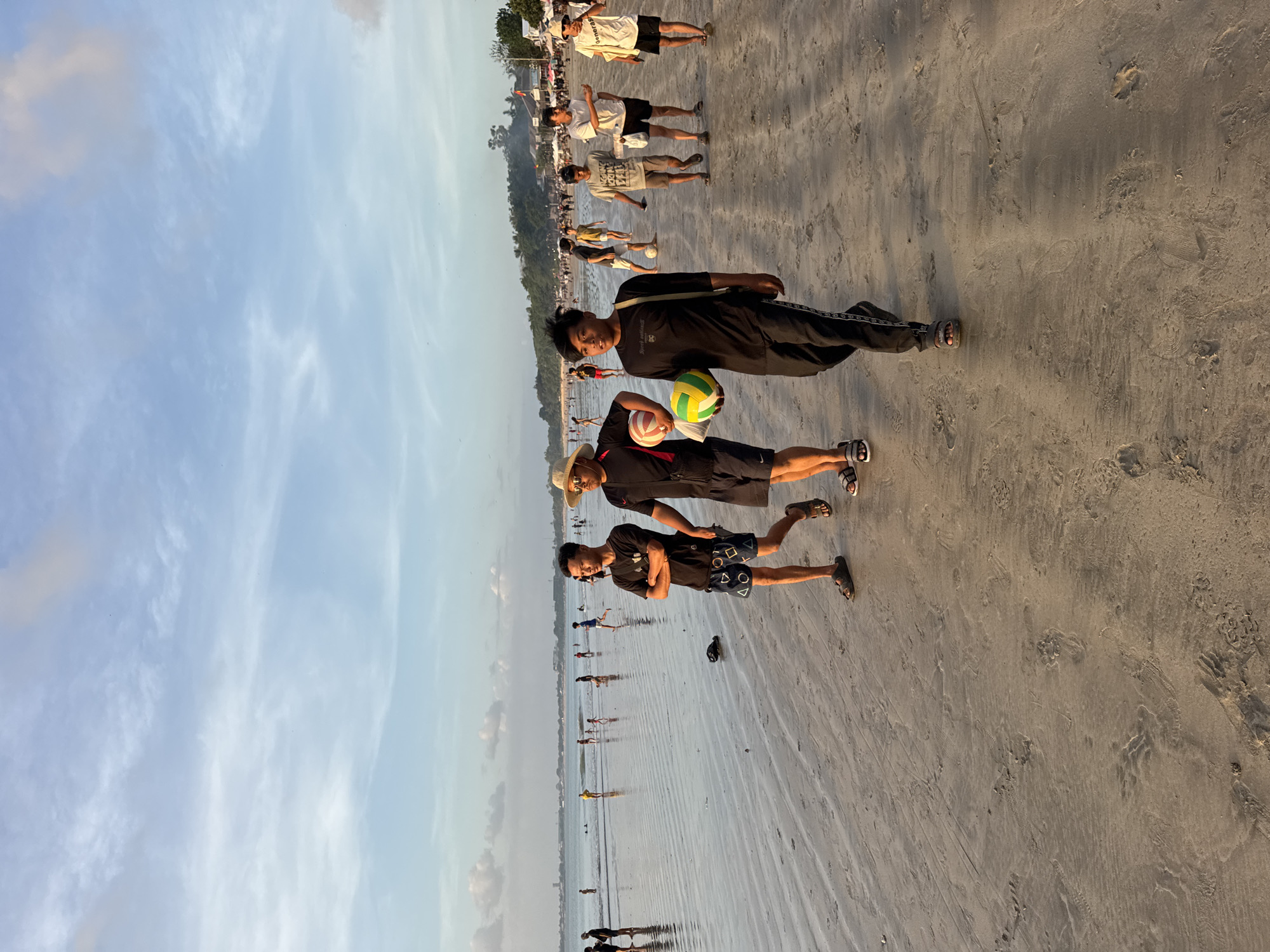 The image shows a beach scene in Kuta Selatan, Indonesia. Three men stand on the sandy beach. One man wears a hat and shorts with a volleyball in his hand. Another man is in the middle wearing shorts. The third man wears a long sleeve shirt and pants. In the background, several other people are visible walking and playing on the beach. The sky is visible with scattered clouds.
