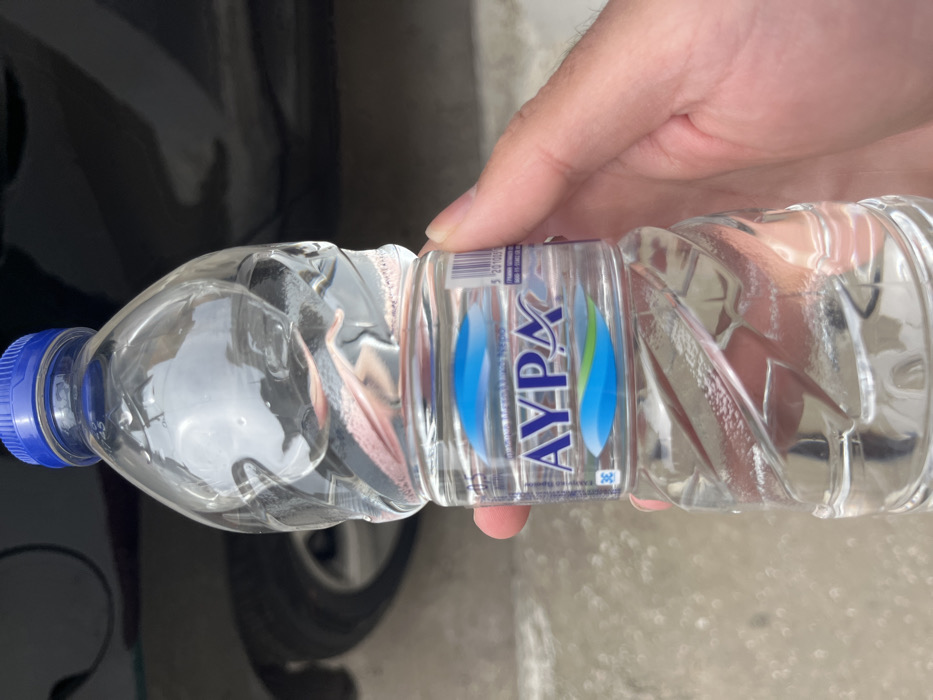 A person is holding a plastic bottle of AYPE water in their right hand. The bottle is mostly full and has a blue cap. The bottle is being held in front of a black car. The scene is set in Limenaria, Greece, at the coordinates latitude 40.627 and longitude 24.575. The pavement is gray and the car is dirty. There are no other people present.