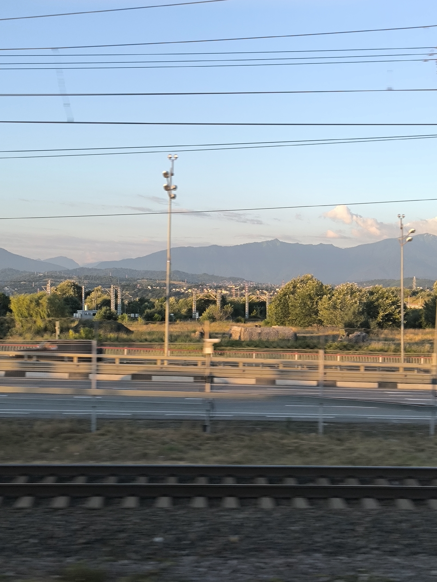 This is an eye-level, long shot, outdoor, horizontal, natural light, daytime photograph taken from a moving train. The scene depicts a mountainous landscape with a city nestled in the valley below, under a clear blue sky with some wispy clouds. In the foreground, there are train tracks and ballast. Above the tracks, there is a road with guardrails and streetlights. Beyond the road, there is a field with some scattered trees, and further in the distance, the city and mountains are visible. There are several power lines running horizontally across the sky. The overall mood is serene and expansive.