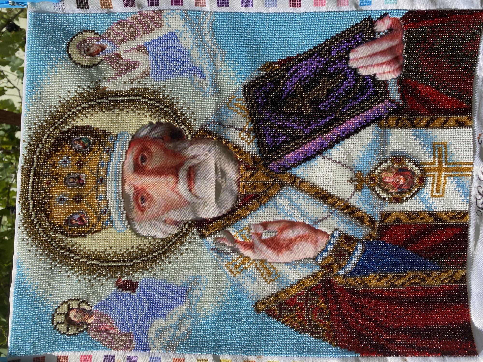 A bead artwork depicts Saint Nicholas, a religious figure, against a light blue background. Saint Nicholas is shown wearing a crown adorned with small religious icons and rich, beaded garments in shades of red, blue, and gold. He holds a book in his left hand and blesses with his right. Flanking him are two figures, possibly angels or other saints, rendered in a similar beaded style, dressed in blue and pink attire. The artwork appears to be hanging outdoors, with glimpses of green foliage and the sky visible in the background. The image is captured from a slightly elevated angle, focusing on the beadwork details. The exact location within Zgrade, Montenegro, is not discernible from the visual information provided in the image.
