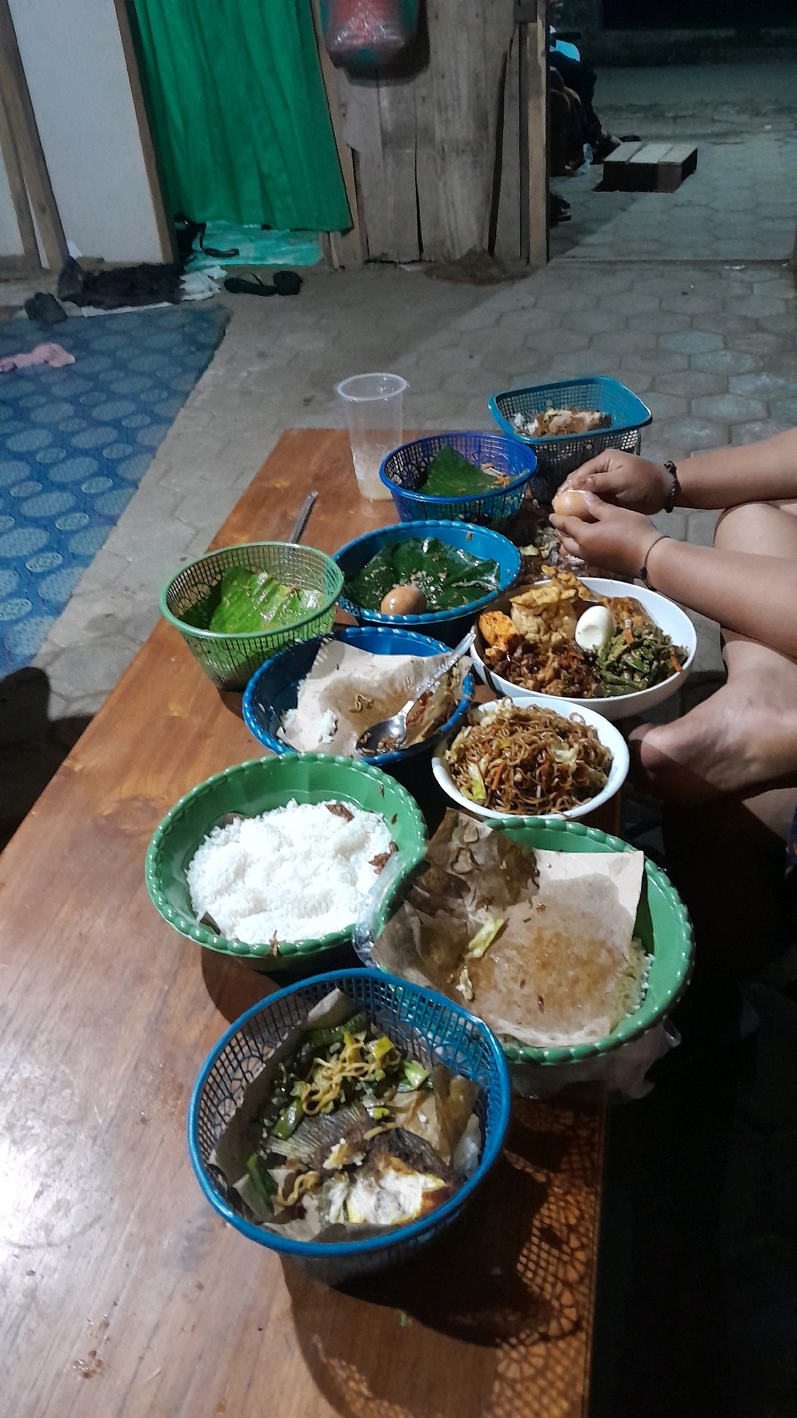 The image depicts an outdoor scene at dusk, likely in Sumberarum, Indonesia, where a group of people are gathered for a meal. A wooden table is laden with various dishes served in colorful woven baskets and bowls. Among the food items are rice, fried noodles, a whole fried fish with accompaniments, and several dishes wrapped in banana leaves, some containing boiled eggs. Two individuals are visible, with one person in the foreground peeling an egg. The background shows a rustic setting with wooden structures and a green curtain, possibly indicating a local eatery or a residential outdoor area. The lighting suggests it is evening, with shadows cast by the objects on the table and floor. The overall atmosphere is casual and communal.