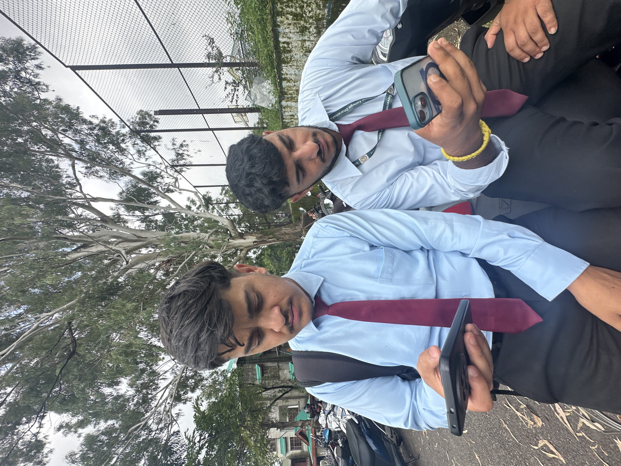 Two young men are sitting outdoors, each holding a smartphone. They are both wearing light blue collared shirts and dark red ties. The man on the left has a black backpack slung over his left shoulder. The man on the right is wearing a yellow bracelet on his left wrist. They are both looking down at their phones, seemingly engrossed in them. The background includes a large tree with sparse leaves, a chain-link fence, and some parked motorcycles. Buildings and greenery are visible in the distance. The overall setting appears to be a campus or public area.