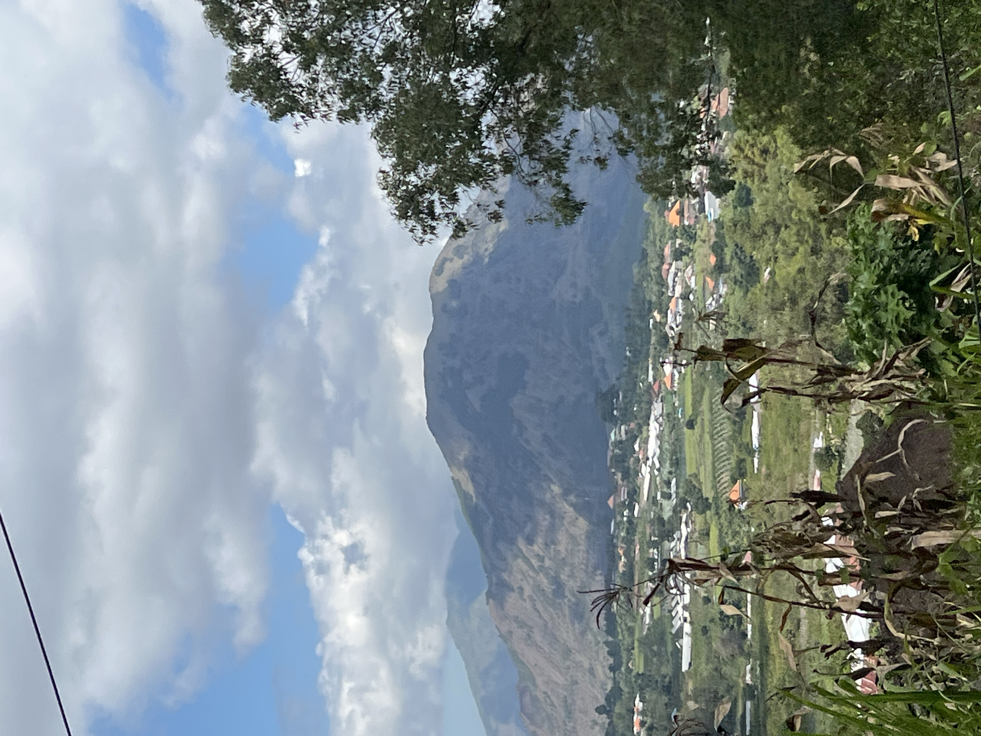 The image is a high-angle view of a village in Kintamani, Indonesia, with a large mountain dominating the background. In the foreground, dry corn stalks and lush green vegetation frame the scene. The village itself is nestled in a valley, characterized by houses with orange-tiled roofs and white walls, interspersed with green fields and trees. The mountain in the background has a rugged, barren appearance, with slopes showing signs of erosion and dry vegetation. The sky above is partly cloudy, with patches of blue sky visible through the white and gray clouds. A thin black wire runs diagonally across the top of the frame. No people are visible in the image.