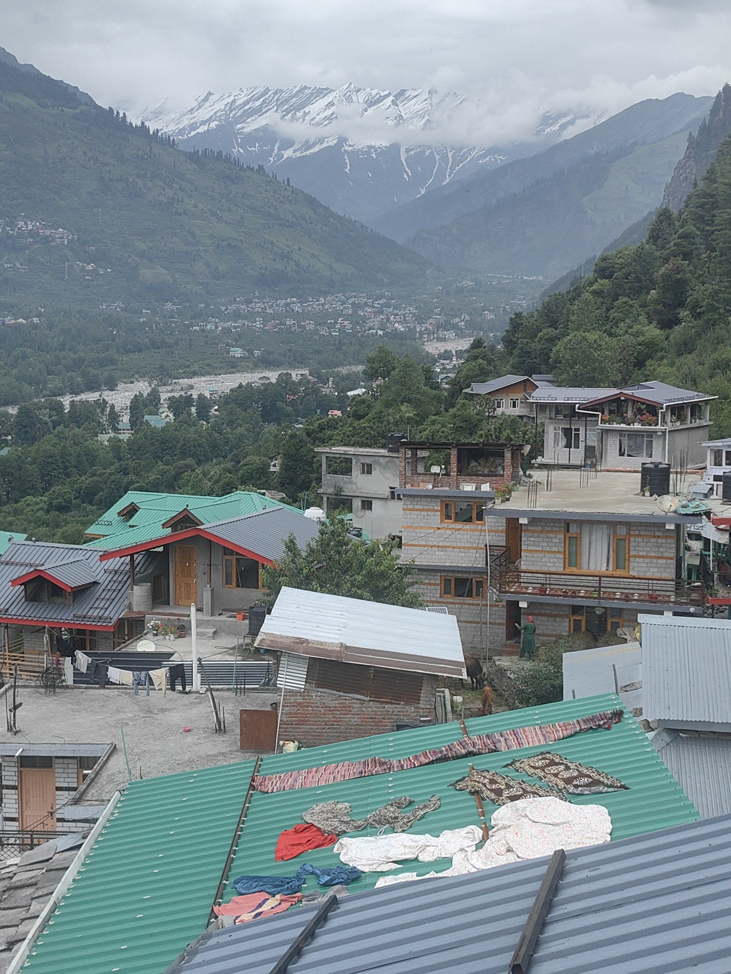 This is a photograph of a village in Vashisht, India, nestled in a valley with snow-capped mountains in the background. The foreground is dominated by the roofs of several buildings, some made of green corrugated metal and others of grey corrugated metal. Clothes are spread out on the green roof to dry, including red, white, and blue garments, as well as patterned fabrics.
The middle ground shows more buildings with varying roof designs, some with red trim. There are also trees and a visible river in the valley. In the distance, more houses and buildings can be seen extending up the mountainside. The sky is overcast with clouds obscuring the mountain peaks. The overall impression is of a densely populated hillside village in a mountainous region.