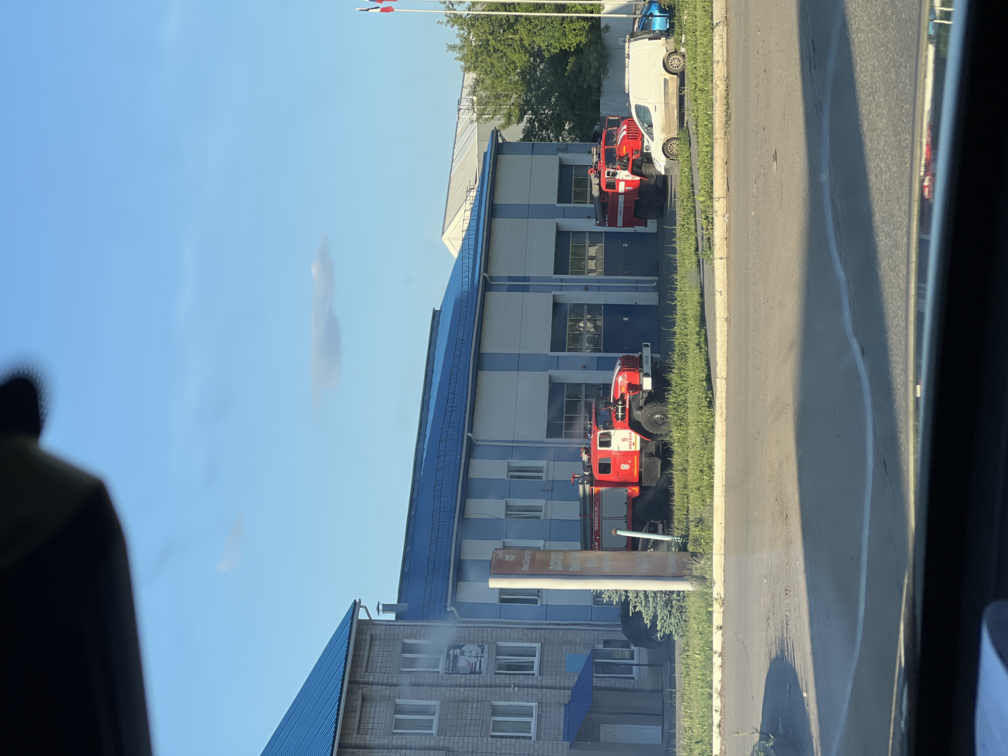 This daytime image, taken from within a vehicle, depicts a street scene in Izhevsk, Russia, under a clear blue sky with minimal cloud cover.
In the midground, two adjacent buildings are visible. The structure on the left is a two-story building constructed of grey brick, featuring multiple rectangular windows and a blue sloped roof. A small blue awning covers an entrance on its ground floor. A poster depicting a person in uniform is affixed to its side. Immediately to its right is a wider building with a facade composed of alternating vertical blue and white stripes, also topped with a blue sloped roof. This building presents at least three large bay doors, characteristic of a garage or vehicle depot.
Two large, red emergency vehicles, identified as fire trucks, are parked directly in front of the striped building. The fire truck on the right clearly displays the Cyrillic inscription "ИЖЕВСК" (Izhevsk) and the number "9" on its side. To the right of this fire truck, a white panel van is parked, facing right, with a blue car partially visible behind it. A dark-colored vehicle is partially visible behind the sign on the left.
A tall, vertical rectangular sign is positioned in the left midground, partially obscuring the left fire truck. Visible Cyrillic text on the sign includes "ДОРОГО" and "ТЕЛЕФОН 01, 101," which are Russian emergency telephone numbers. A small evergreen tree is planted near the base of this sign.
On the far right, two flagpoles stand erect. The uppermost flag is identifiable as the Russian national flag, displaying horizontal white, blue, and red stripes. The lower flag also exhibits a similar tri-color pattern.
A paved road occupies the immediate foreground, separated from the buildings by a strip of green grass. Shadows on the road indicate direct sunlight.