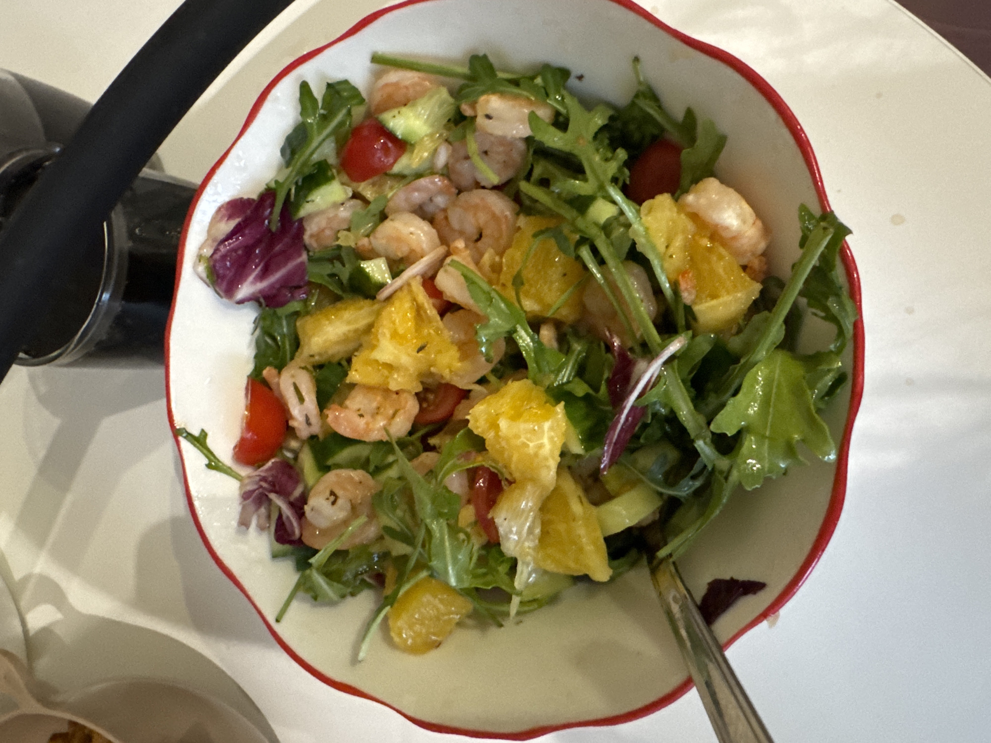 A top-down shot shows a white bowl with a red rim, filled with a shrimp and orange salad. The salad consists of shrimp, orange segments, arugula, cucumber slices, red tomato pieces, and red lettuce leaves. A silver spoon rests on the right side of the bowl. To the left of the bowl, partially visible, is a glass of dark liquid and a white surface. The scene is a table setting, likely indoors. The location is not discernible from the image, and there is no evidence to suggest the location is Chaikovsky, Russia.