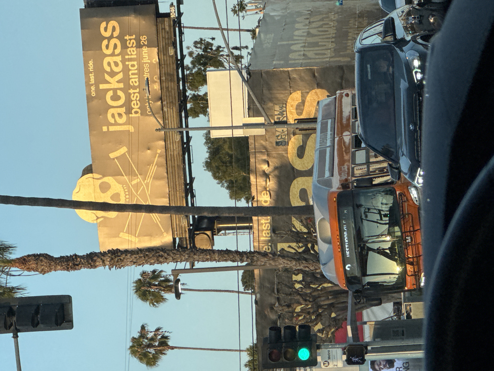 The image captures a street scene in Los Angeles, California, with a prominent billboard advertising "Jackass: Best and Last" with a release date of June 26. The billboard features a skull and crossbones motif with crutches. In the foreground, an orange and white Metro bus is visible, along with a dark-colored SUV. Traffic lights, palm trees, and a clear blue sky contribute to the urban, outdoor setting. The overall lighting suggests it is daytime, likely late afternoon or early evening given the golden hue of the sunlight.