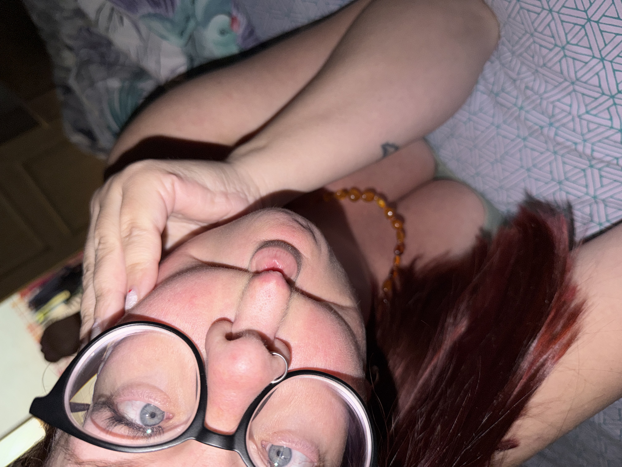 A close-up, upside-down selfie shows a person wearing black-rimmed glasses and a nose ring. The person is lying down on a bed with patterned bedding. An amber beaded necklace is visible around the person's neck. Brownish-red hair is visible in the lower right corner. A hand is near the person's forehead. The location is unknown and is not identifiable from the provided image. The city of Chaikovsky, Russia, is not discernible from the image.
