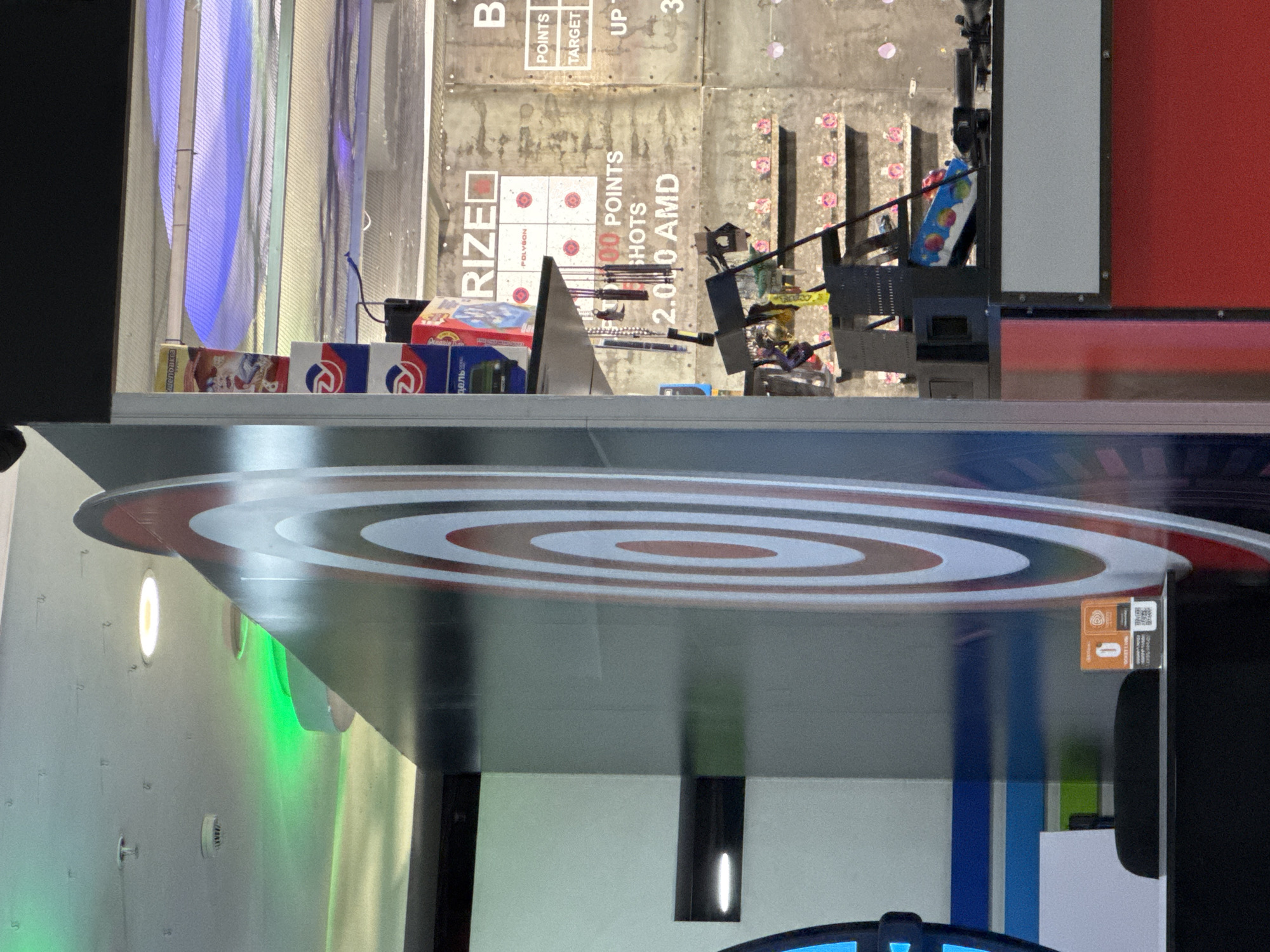 This image captures an indoor archery or target shooting range, likely within an entertainment or gaming center in Yerevan, Armenia. The original image is rotated 90 degrees clockwise.
In the background, a textured grey wall serves as the target area, marked with grids for scoring. Various square targets, each featuring a red bullseye, are visible. Some targets are labeled "RIZE" and "POLYGON". Text on the wall includes "POINTS", "TARGET", "SHOTS", "UP 3", "000 POINTS", and "2000 AMD", indicating a game or activity with a scoring system and a cost in Armenian Dram. Several arrows rest in a bow stand or holder on the ground in front of these targets. Behind the stand, a blue and white patterned bag or object is discernible.
The foreground features a large, reflective surface with a prominent circular target design in red, white, and grey concentric rings. This target is likely part of the shooting setup. The surface reflects some of the background elements. To the left side, several boxes are stacked; some bear a stylized "R2" logo, while others have Cyrillic text, notably "ЖЕЛЕЗНОЕ ДЕЛО" (ZHELEZNOE DELO), which translates to "Iron Business" or "Hard Fact".
Artificial lighting illuminates the scene, with distinct green and blue glows visible in the reflections and on the walls. A small orange label with the number "0", a QR code, and text including "Polygun" and "rize.am" is visible on a stand or monitor to the right. No people are present in the image.