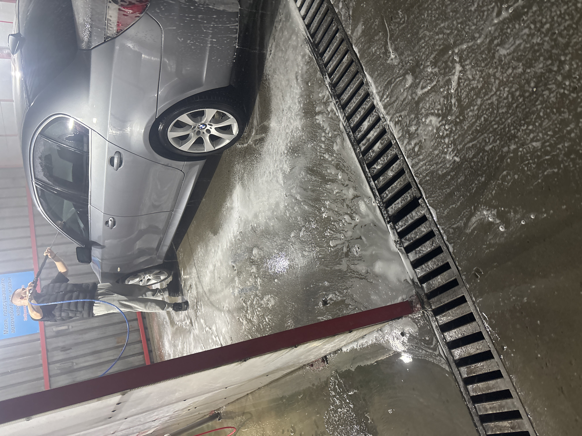 A man washes a silver BMW sedan at a car wash. The car is partially visible, with the front passenger side facing the viewer. The man is holding a pressure washer wand, directing the spray towards the car. The ground is wet and covered in soapy water. A drainage grate runs along the edge of the washing bay. The scene takes place in a car wash in the city of Plovdiv, Bulgaria, evidenced by the Cyrillic text on the wall.