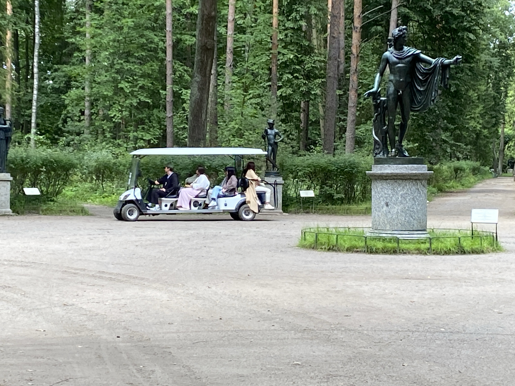 A group of people ride in a white golf cart through a park with tall trees. A dark bronze statue of a male figure stands on a stone pedestal on the right side of the image. The golf cart has a white canopy and is moving from left to right. There are four people visible in the golf cart. The driver is a man wearing a dark shirt and hat. The passengers are two women in light-colored clothing and one person in a tan outfit. The ground is a dirt or gravel path. The environment appears to be a park or garden with lush green trees and bushes. There is a small fence around the base of the statue. The scene suggests a recreational activity in a historical or natural setting, possibly within the Pavlovsk Palace and Garden Complex in Pavlovsk, Russia, which is known for its extensive parklands and classical sculptures.