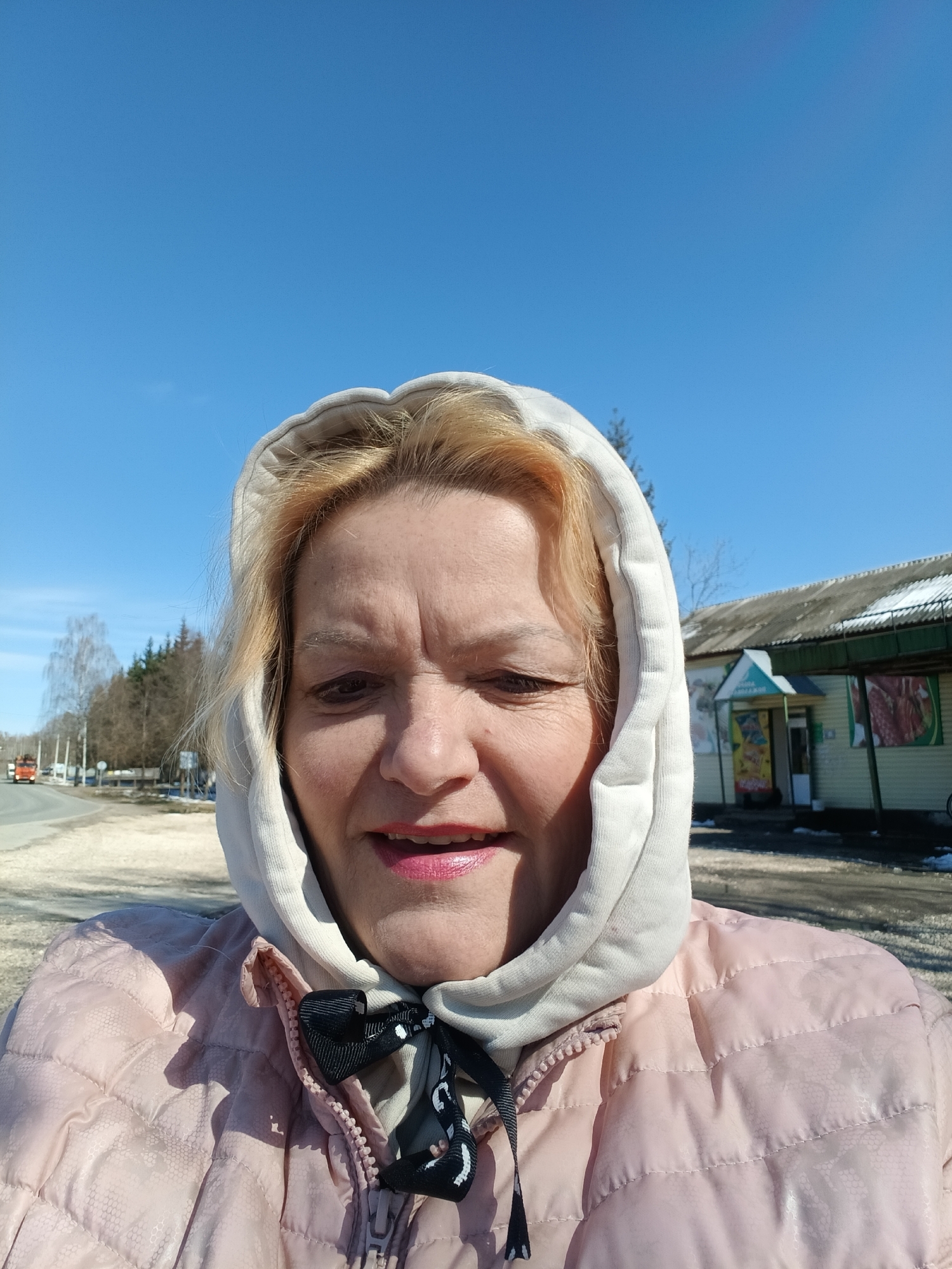 A medium close-up captures a woman in Красный Маяк, Russia. She wears a light pink puffer jacket and a white hooded top with a black and white patterned tie at the neck. Her blonde hair is visible under the hood. The background shows a clear blue sky and a road. In the distance, there is a building with a green awning and advertisements visible on its exterior. Trees are sparsely scattered along the roadside.