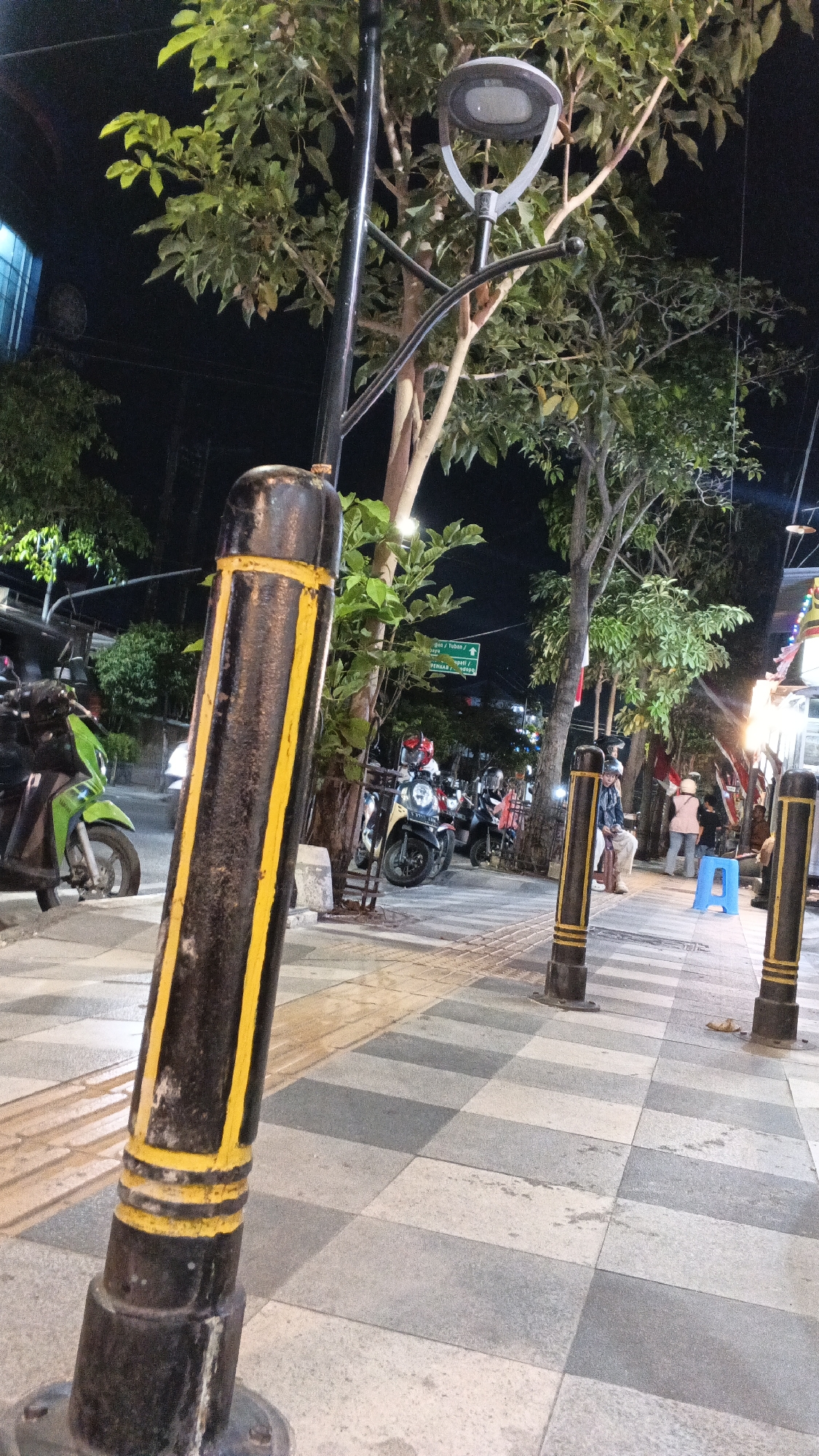 The image depicts a street scene at night in Bojonegoro, Indonesia, likely on a pedestrian walkway adjacent to a road. In the foreground, a black bollard with yellow stripes is prominently visible, positioned on a checkered patterned sidewalk made of large grey and white tiles. To the left, a bright green scooter is parked, and further down the street, several other motorcycles are parked in a row. Trees with green foliage line the sidewalk, and streetlights illuminate the area. In the background, there are buildings with some lights on and a green street sign pointing towards "Gresik / Tuban / Lamongan" and "Cepu / Ngawi". A few people are visible on the sidewalk, some standing and others walking. The overall atmosphere suggests a public area in a city at night, possibly a market street or a busy thoroughfare.