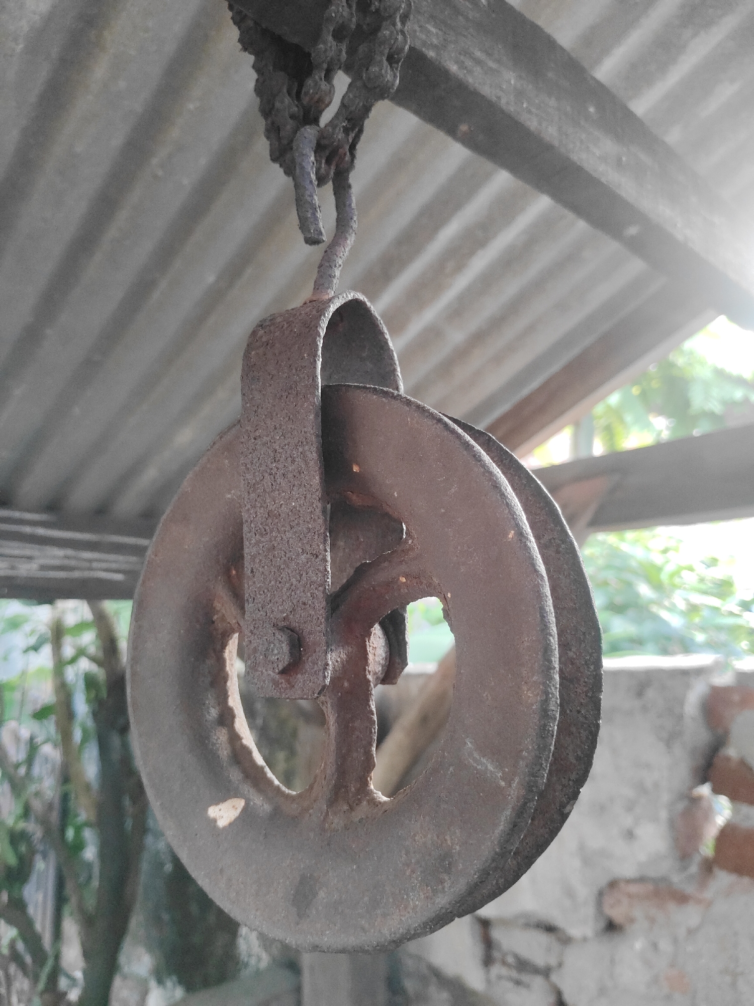 An aged, rusty pulley hangs from a chain, suspended beneath a corrugated metal roof supported by wooden beams. The pulley, made of metal, shows signs of significant wear and rust, with two large wheels designed to guide a rope or chain. The backdrop includes hints of greenery and a brick wall, suggesting an outdoor setting. The lighting indicates it might be daytime, with some sunlight filtering through. The image's composition focuses on the pulley and its immediate surroundings. The specific location in Surabaya, Indonesia, is not visually discernible from the image itself.
