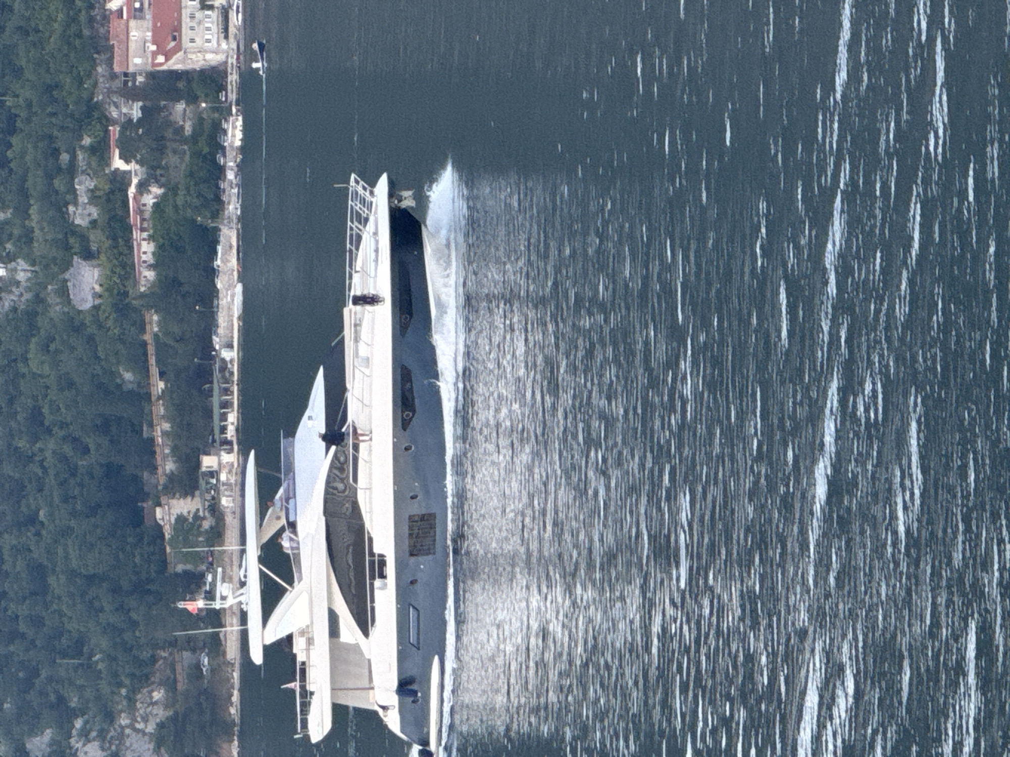 Aerial view of a large yacht moving through water, creating a visible wake. The vessel is passing by the shoreline of Đurići, Montenegro, where buildings are visible along the coast. Vegetation covers the land.
