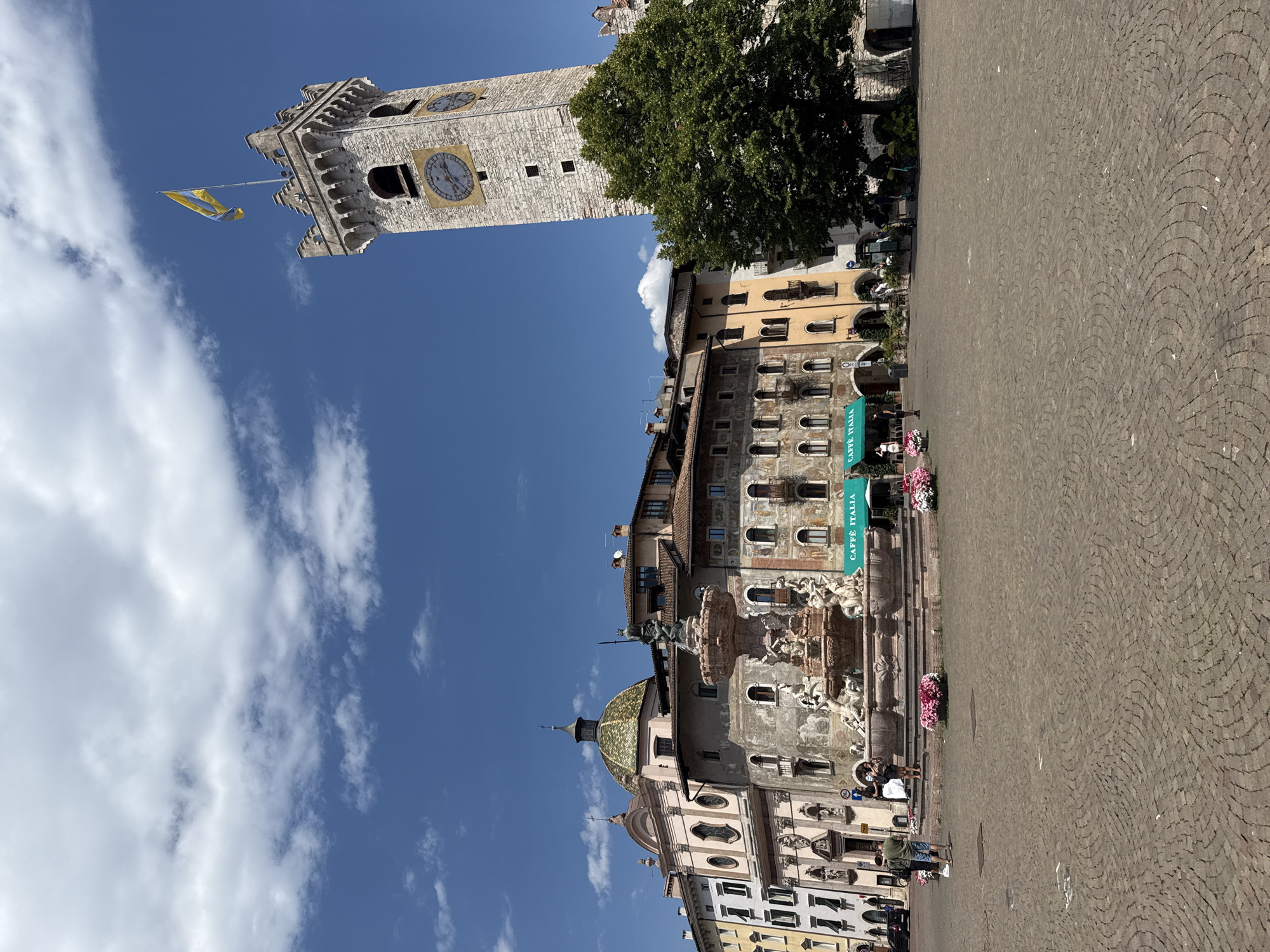 This image depicts a sunny daytime scene in a European city square. Dominating the sky is a tall, stone clock tower with a yellow and blue flag flying from its side. Below the tower, a series of buildings with arched windows line the square, some adorned with intricate architectural details. A large green tree partially obscures one of the buildings.
In the foreground, a cobblestone plaza stretches out, with a few people visible walking or sitting. A decorative fountain with sculptures is a central feature of the square. A green banner with the text "CAFFÈ ITALIA" hangs from one of the buildings, indicating a cafe. Pink flowers are placed in planters near the fountain and cafe. The sky is a clear blue with scattered white clouds, suggesting pleasant weather. The overall impression is one of a historical European town square on a bright day.