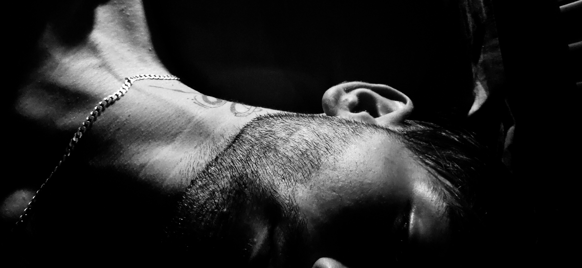 The black and white, close-up, waist-up photograph shows a man's face and neck in profile, set against a dark background. The lighting casts a strong shadow across the right side of his face, obscuring his eye and nose. His left ear is visible, along with stubble on his cheek and jawline. A silver chain necklace rests on his collarbone, and a tattoo of concentric circles is visible on his neck. The image is grainy, suggesting a low-light environment or a stylistic choice. The location is unknown.