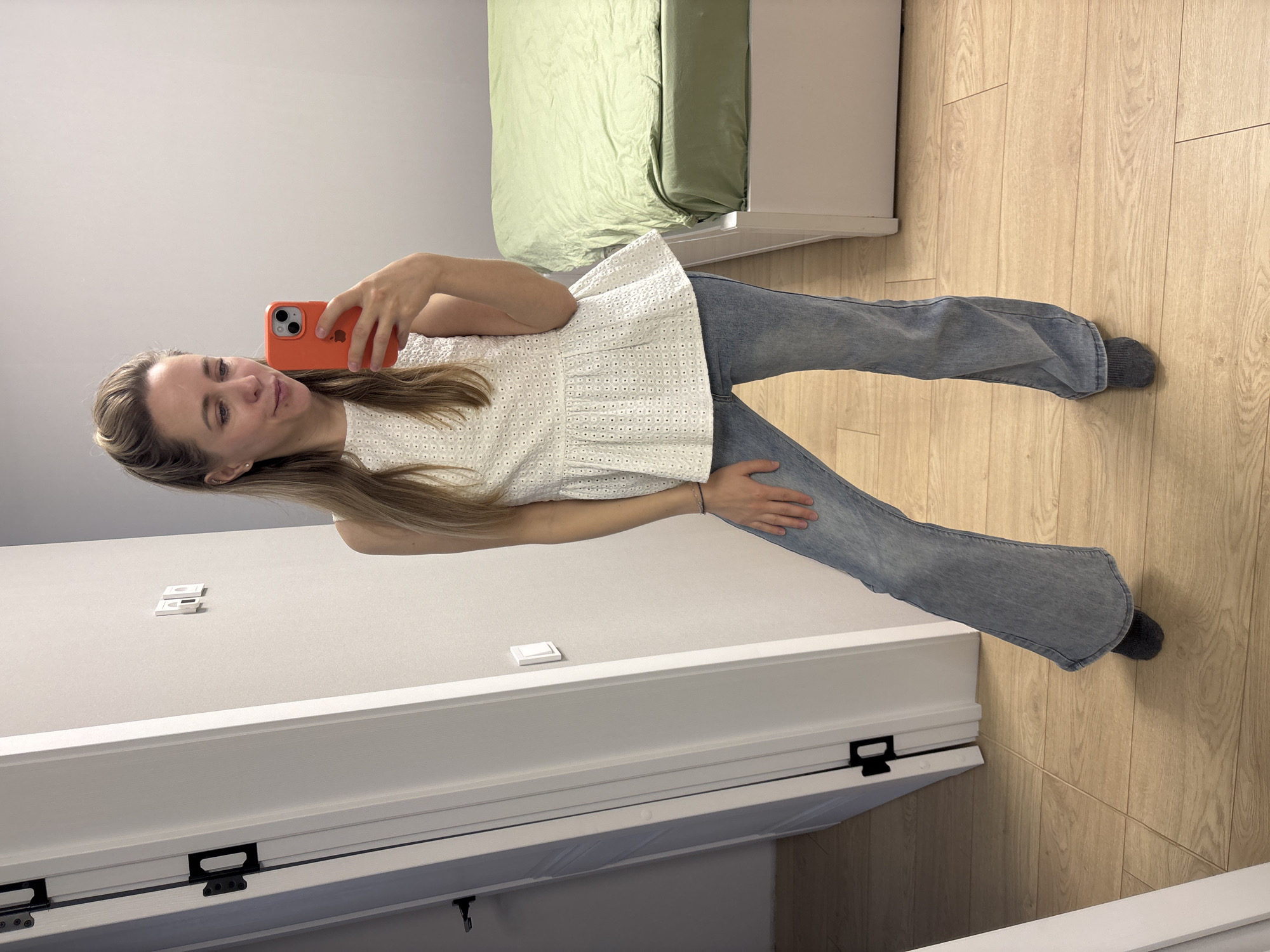 A young woman takes a mirror selfie indoors, captured in a horizontally rotated image. She has long, light brown hair and a pleasant, subtle smile. She is wearing a white, eyelet-embroidered peplum top and light blue, flared jeans, with dark grey socks visible on her feet. In her right hand, she holds a bright orange-cased iPhone, displaying a visible Apple logo on its back, while her left hand rests on her hip.
The setting is a modern indoor room, likely in Chaikovsky, Russia, featuring plain light grey walls and light wood-plank flooring. In the background, a bed with light green bedding is visible. The foreground shows a white door or cabinet with black hardware. The room is brightly lit.