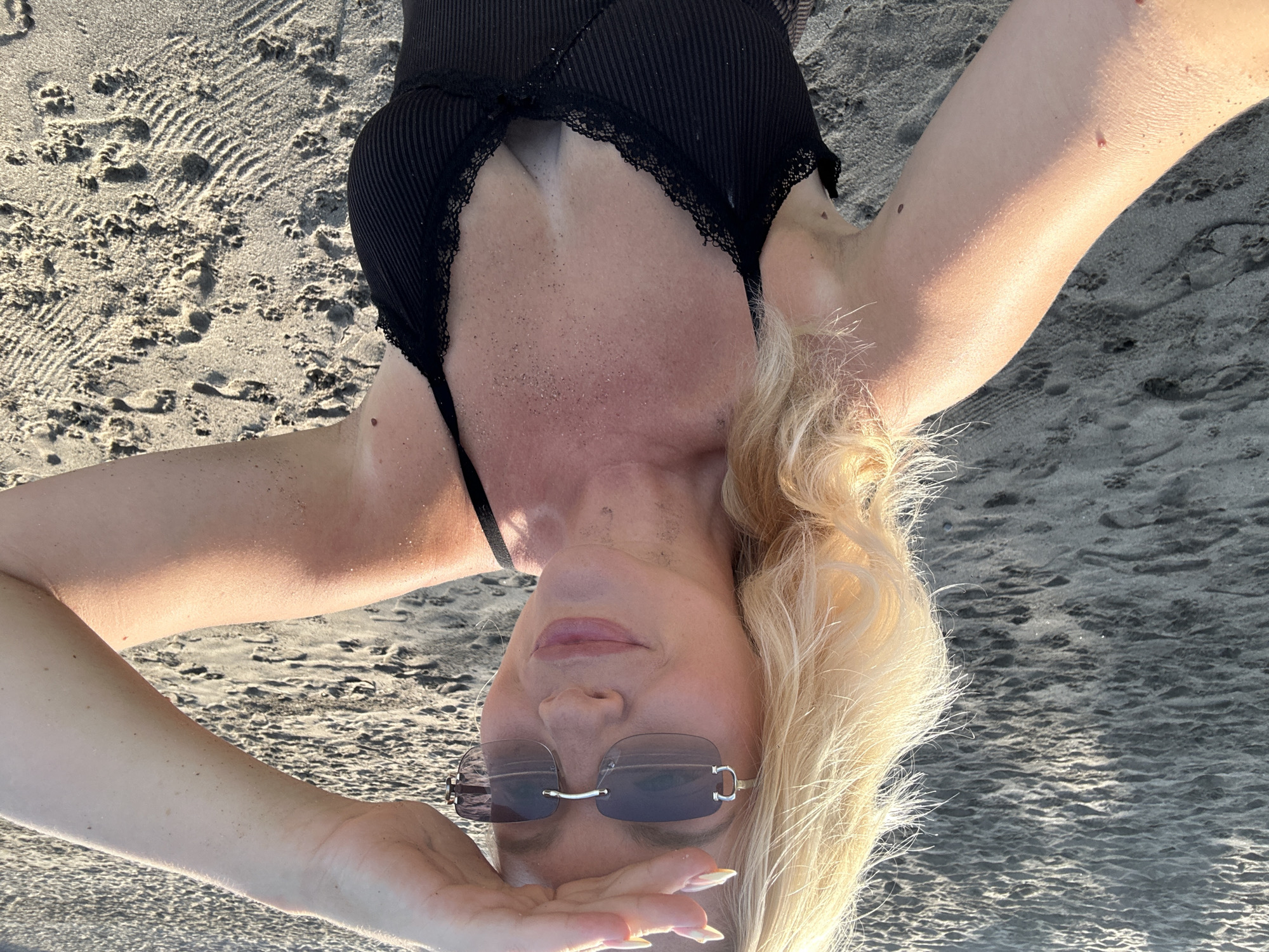 A blonde woman with fair skin is taking a selfie on a sandy beach. She is wearing a black swimsuit with lace trim and sunglasses. She is shielding her eyes from the sun with her right hand. The sand is light brown and has footprints and ripples from the tide. The lighting suggests it is late afternoon or early morning. The background is out of focus, showing more sand. The location is identified as North Kuta, Indonesia.