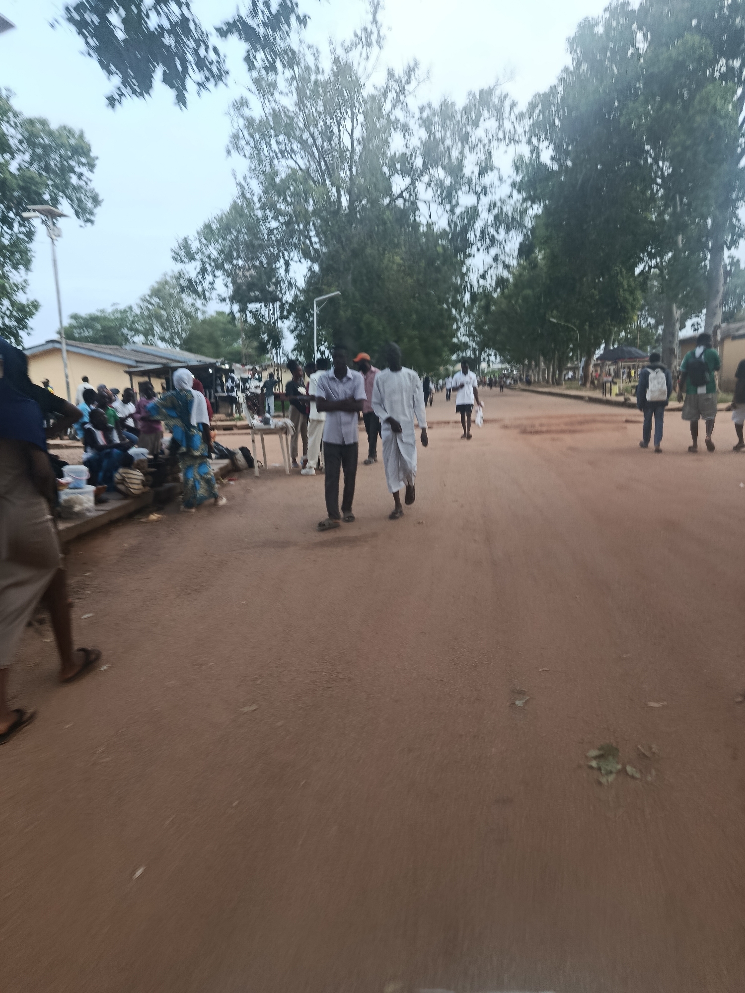 The image depicts a street scene in Rafin Par, Nigeria, with a reddish-brown dirt road and scattered vegetation. Several people are visible, some walking along the road and others gathered on the left side of the image, seated on a raised concrete structure.
In the foreground, a person wearing a blue headscarf and a brown skirt walks away from the camera. Two men in traditional attire are walking towards the camera in the middle of the road; one is wearing a white robe and the other a light-colored shirt and trousers. On the left, a group of people are seated, with one woman in a blue and white patterned dress standing. There are also individuals with backpacks walking further down the road. Buildings with light-colored walls are visible in the background, along with trees and streetlights. The lighting suggests it is either early morning or late afternoon.