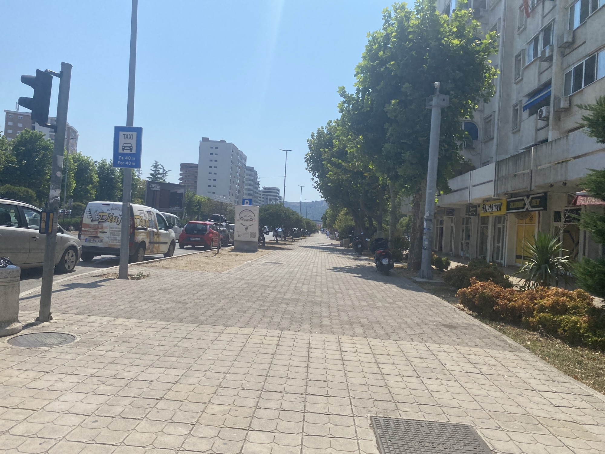 The image shows a street scene in the city of Bar, Montenegro, on a clear, sunny day. A wide pedestrian walkway paved with interlocking concrete tiles runs along the right side of the frame. On the left side of the walkway, there is a traffic light, a lamppost with a blue taxi sign, and several parked vehicles, including a white van and a red car. Several trees with dense green foliage are planted along the left side of the walkway, casting shadows onto the pavement. In the background, multi-story buildings with light-colored facades and numerous windows are visible. Further down the street, more trees and buildings can be seen. A few motorcycles are parked on the right side of the walkway, near the edge of the road. The overall scene suggests a commercial or residential area within the city.