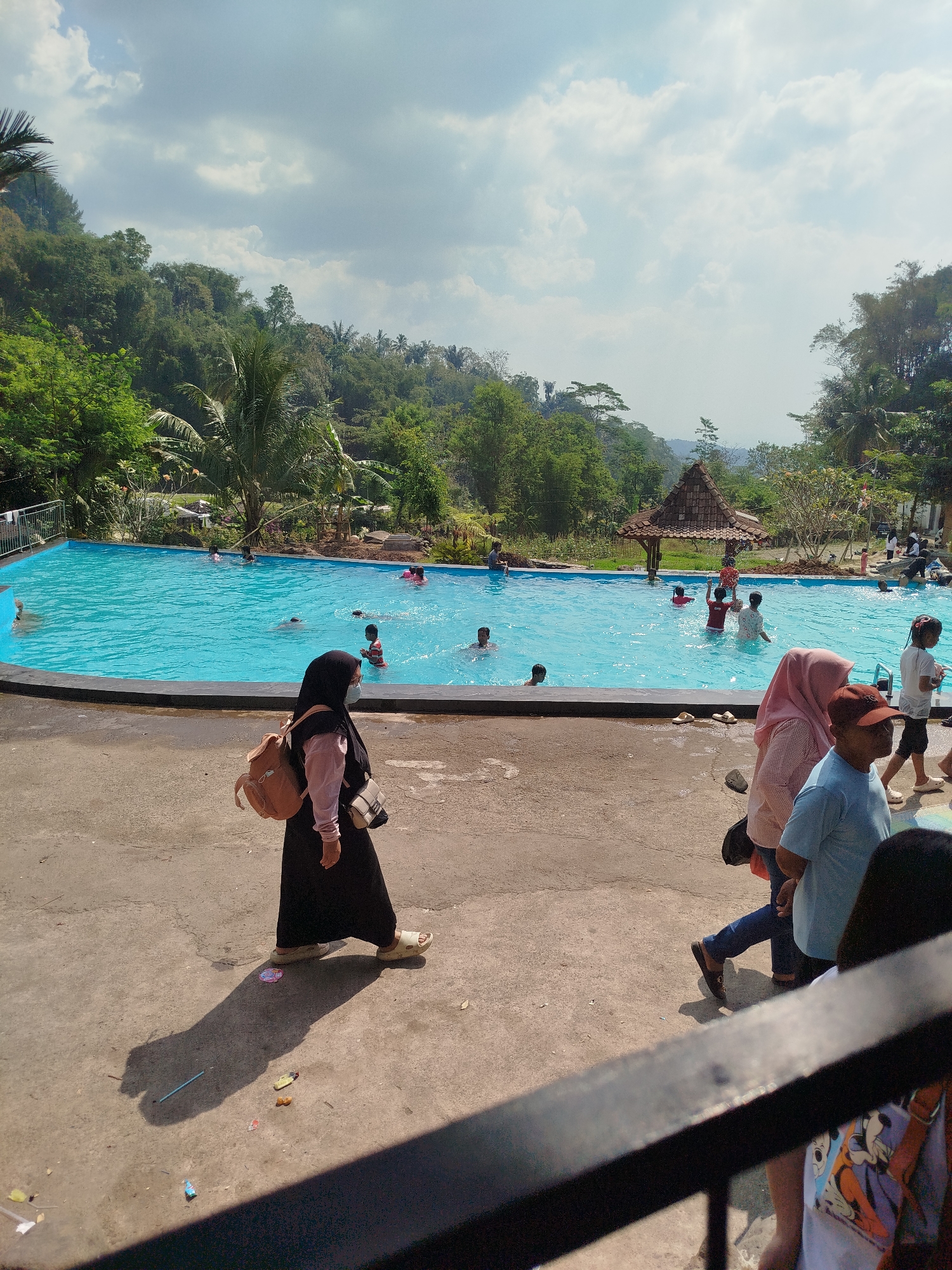 This is a full-shot, eye-level, outdoor photograph taken on a sunny day in Ngesrepbalong, Indonesia. The image captures a bustling public swimming pool set against a backdrop of lush, green hills. In the foreground, several individuals are walking on a concrete path, some in casual attire, while others are dressed modestly with head coverings. One woman in a black hijab and pink shirt is walking away from the camera, carrying a light brown backpack. Further to the right, another woman in a pink hijab and a man in a blue t-shirt and brown hat are also walking.
The middle ground is dominated by a large, bright blue swimming pool filled with water. Numerous people, including children, are enjoying the pool, with some swimming and others playing in the water. A traditional-style gazebo with a thatched roof is situated near the edge of the pool, providing shade.
The background features a dense forest that covers the rolling hills, with palm trees and other tropical vegetation visible. The sky is partly cloudy, with white cumulus clouds scattered across a light blue expanse. The overall scene depicts a lively recreational area within a natural, scenic environment.