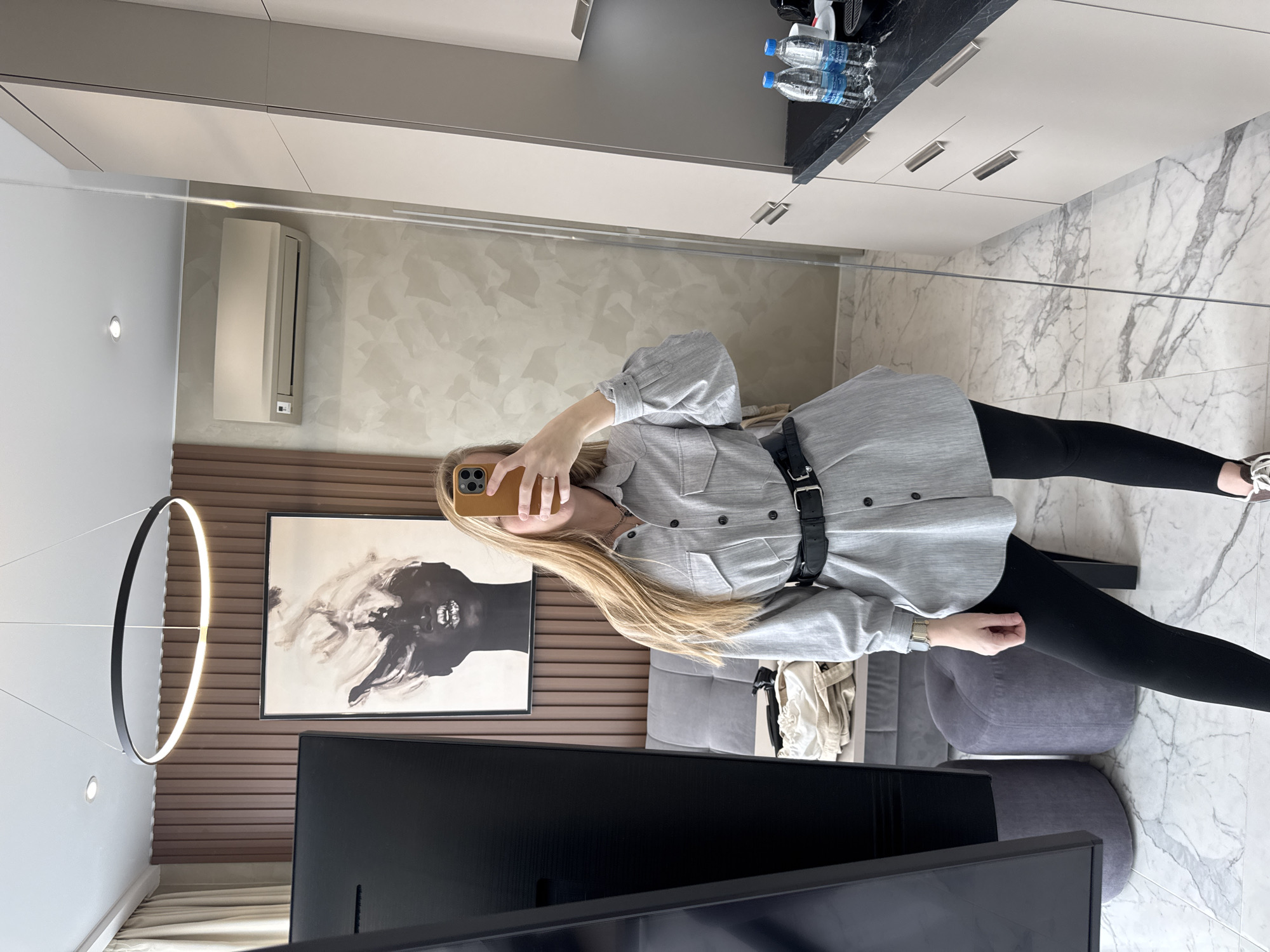 A woman stands in a room, likely an apartment or hotel room, taking a picture of herself in a mirror. She is wearing a grey button-down shirt, black leggings, and a wide black belt. She is holding a phone with a gold-colored case in her left hand, obscuring her face. Her long blonde hair is visible.
The room features modern interior design. There's a flat-screen television in the foreground and a grey sofa behind it. A piece of artwork depicting a face in black and white hangs on a horizontally slatted wall behind the sofa. A circular, modern light fixture hangs near the artwork. An air conditioning unit is mounted on a wall. The floor is marble-patterned tile. The kitchen area is visible, with light cabinets and a dark countertop. Bottles of water are placed on the countertop. The location is likely Sochi, Russia.