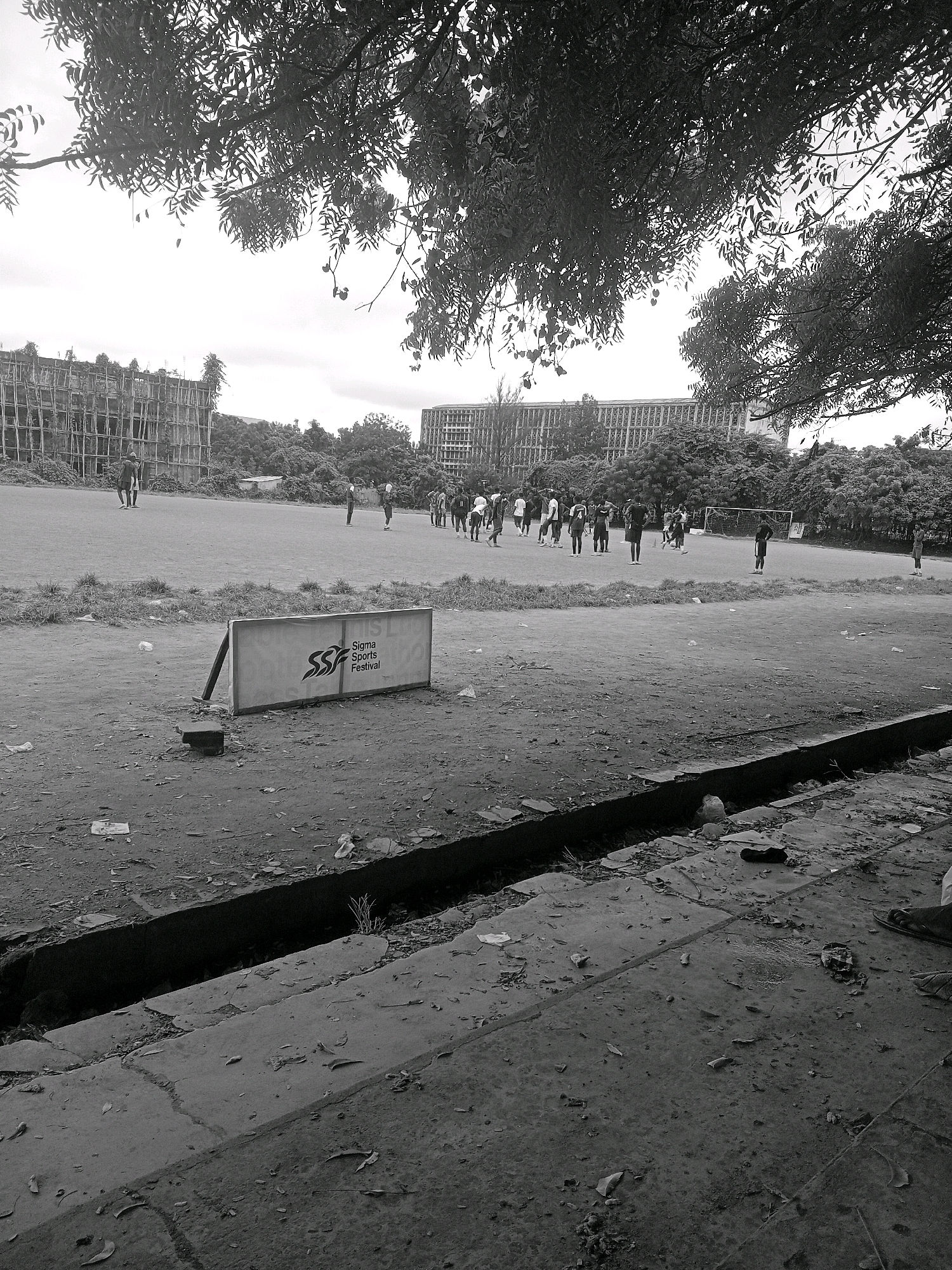 This is a grayscale, medium-angle, outdoor, daytime photograph of a sports field in Ibadan, Nigeria, likely on a university campus given the presence of large, multi-story buildings. The foreground features a cracked concrete drainage ditch filled with debris, and a concrete walkway with scattered leaves and litter. A white sign with the text "Sigma Sports Festival" is propped up on a dirt patch in the middle ground, to the left. Beyond the sign, a large, open, grassy sports field is visible. Several groups of people, predominantly young adults, are scattered across the field. One group of approximately ten individuals is gathered near the center, appearing to be engaged in some form of athletic activity or waiting for instructions. Further in the distance, another group of about five people are positioned near a soccer goal. Two tall, unfinished or derelict buildings with exposed scaffolding and concrete structures are visible on the left side of the frame, while a long, multi-story building with rows of windows occupies the background behind the sports field. Trees with dense foliage line the perimeter of the field and the background. The sky is overcast, creating a diffused light.