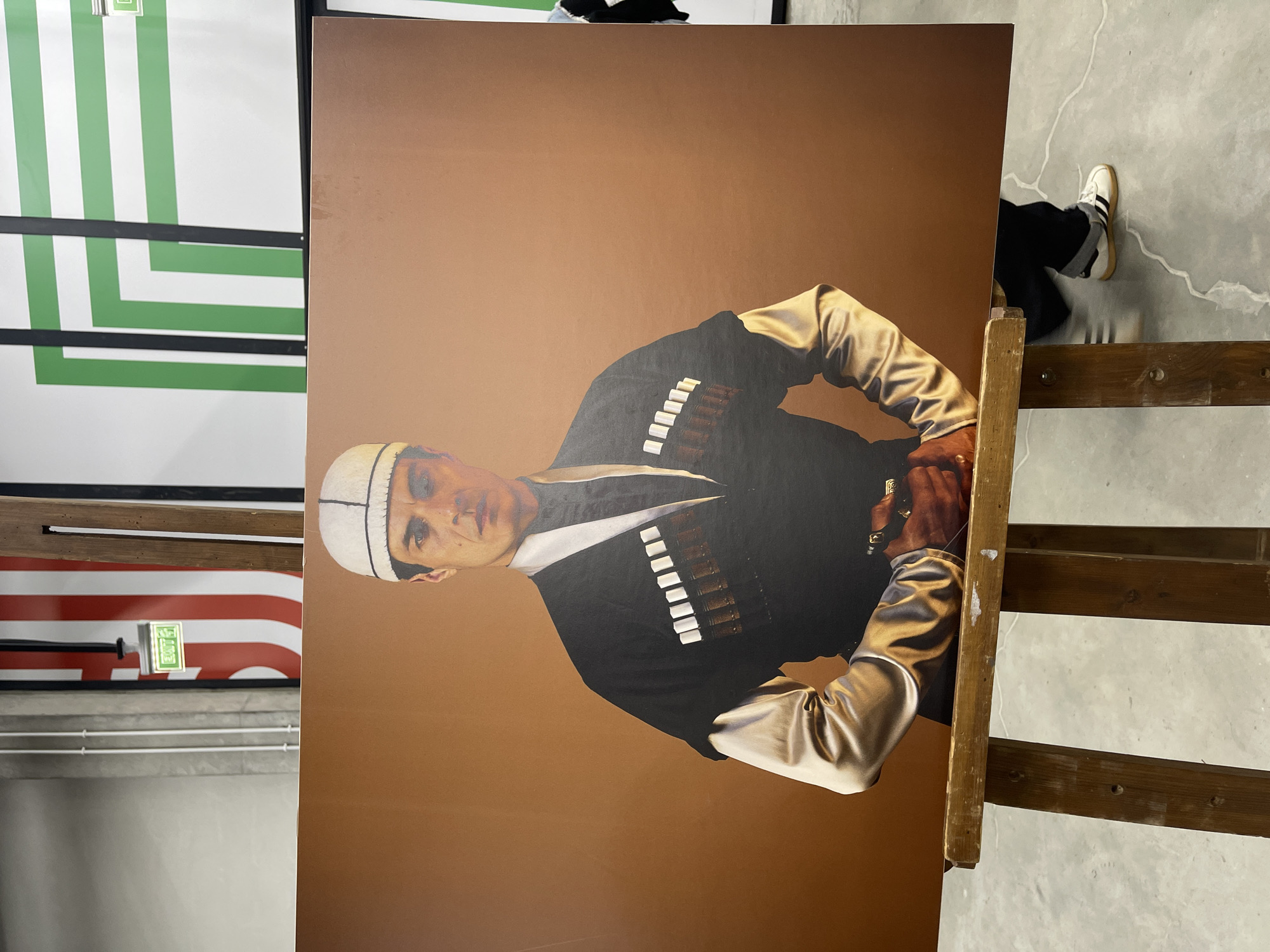 The image depicts a large portrait propped up on a wooden easel, likely in a gallery or exhibition space in Batumi, Georgia. The portrait features a man in traditional Georgian attire, including a chokha (a high-necked woolen coat adorned with decorative cartridge pockets) and a papakha (a tall fur hat). The background of the portrait is a solid brown color. The man has fair skin, dark hair, and is looking towards the viewer.
To the left of the portrait is a display featuring alternating green and white stripes, framed in black. Below this is another display, showcasing red and white stripes, reminiscent of the American flag. An illuminated exit sign is visible on this lower display.
The floor appears to be concrete. In the background, behind the easel, a person wearing black pants and white sneakers can be partially seen. The easel is made of unfinished wood. The setting suggests an art exhibition or gallery space with contemporary displays and the portrait as a featured artwork.