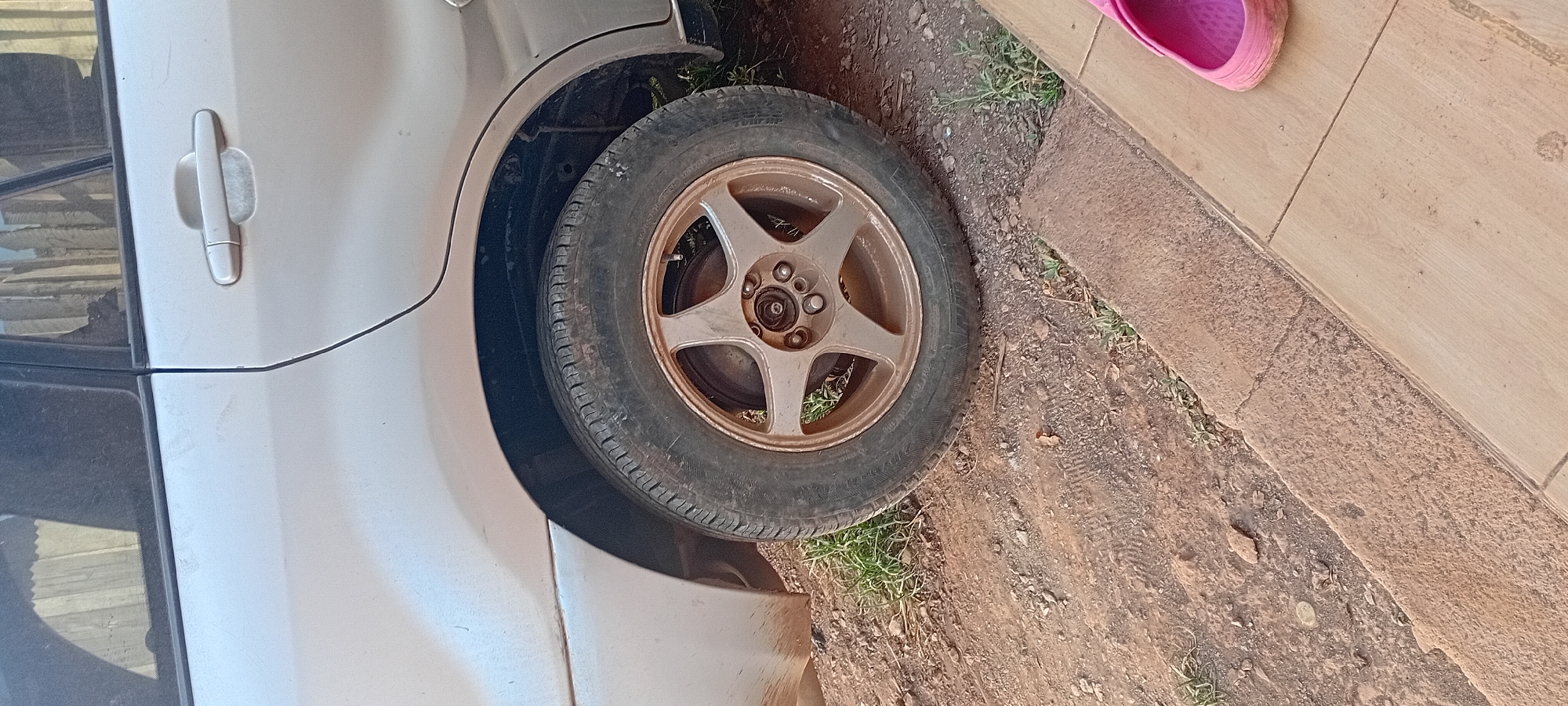 A side view of the rear wheel of a silver vehicle is shown. The tire is slightly worn, and the rim is a metallic bronze color with five spokes. The vehicle is parked on a dirt and gravel surface with some patches of grass. A concrete curb runs along the right side of the image, separating the dirt area from a paved walkway. A single pink sandal is visible on the paved walkway in the lower right corner of the frame. The background shows a vertical striped surface, possibly a fence or corrugated metal sheeting. The scene appears to be outdoors, possibly in a residential or parking area in Kijabe Ward, Kenya.