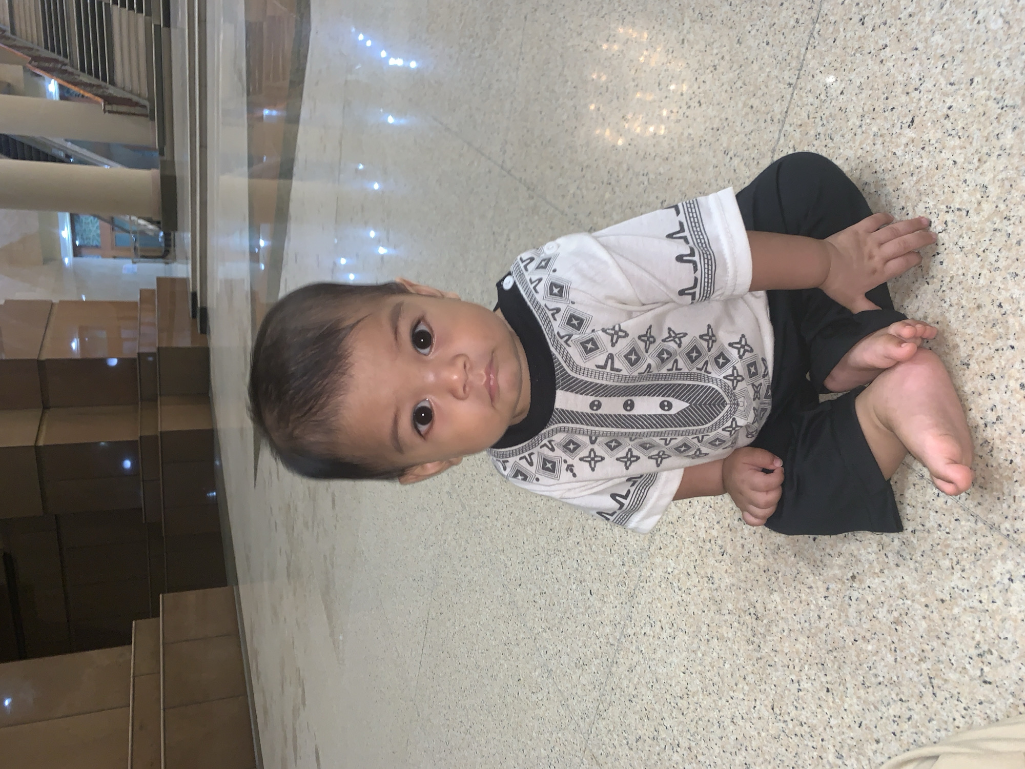A baby is depicted sitting upright on a light-colored, speckled floor, looking directly at the camera. The baby has dark hair and dark eyes, and appears to be of Asian descent. He is wearing a white short-sleeved shirt with black and grey geometric patterns around the collar, sleeves, and down the front, paired with dark-colored pants. His hands are resting on his legs, and his bare feet are visible.
The setting appears to be indoors, likely within a building in Samarinda, Indonesia. The floor has a polished, reflective surface, showing bright, scattered light reflections. In the background, there are large, light-colored pillars and a set of stairs made of similar material, suggesting a spacious hall, lobby, or a place of worship. Further in the background, a dark wooden door or window with a patterned grate is visible. The lighting suggests it is daytime or a brightly lit interior. The baby's expression is neutral and observant. No discernible text is visible in the image.