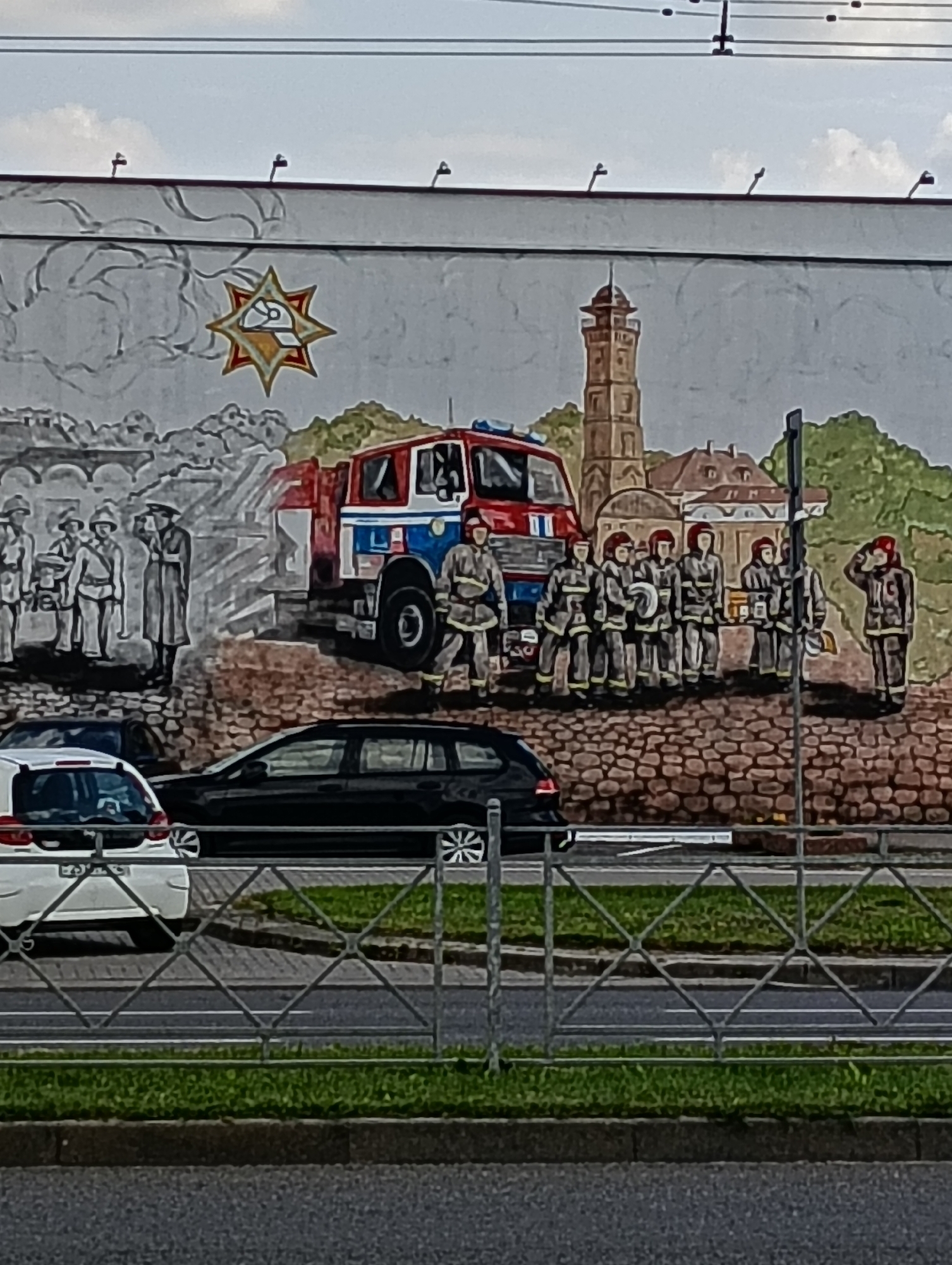 The image depicts a mural on a building wall in Hrodna, Belarus, showing firefighters and a fire truck. The mural features a group of firefighters in uniform, some standing with equipment and others in a line. A red fire truck is prominently displayed in the center. Behind the firefighters, there is a cityscape with buildings and a tower, rendered in a muted color palette with some elements in grayscale. A distinctive star symbol with a cap in the center is depicted to the left of the firefighters. The mural is situated above a street with cars and a metal fence in the foreground.