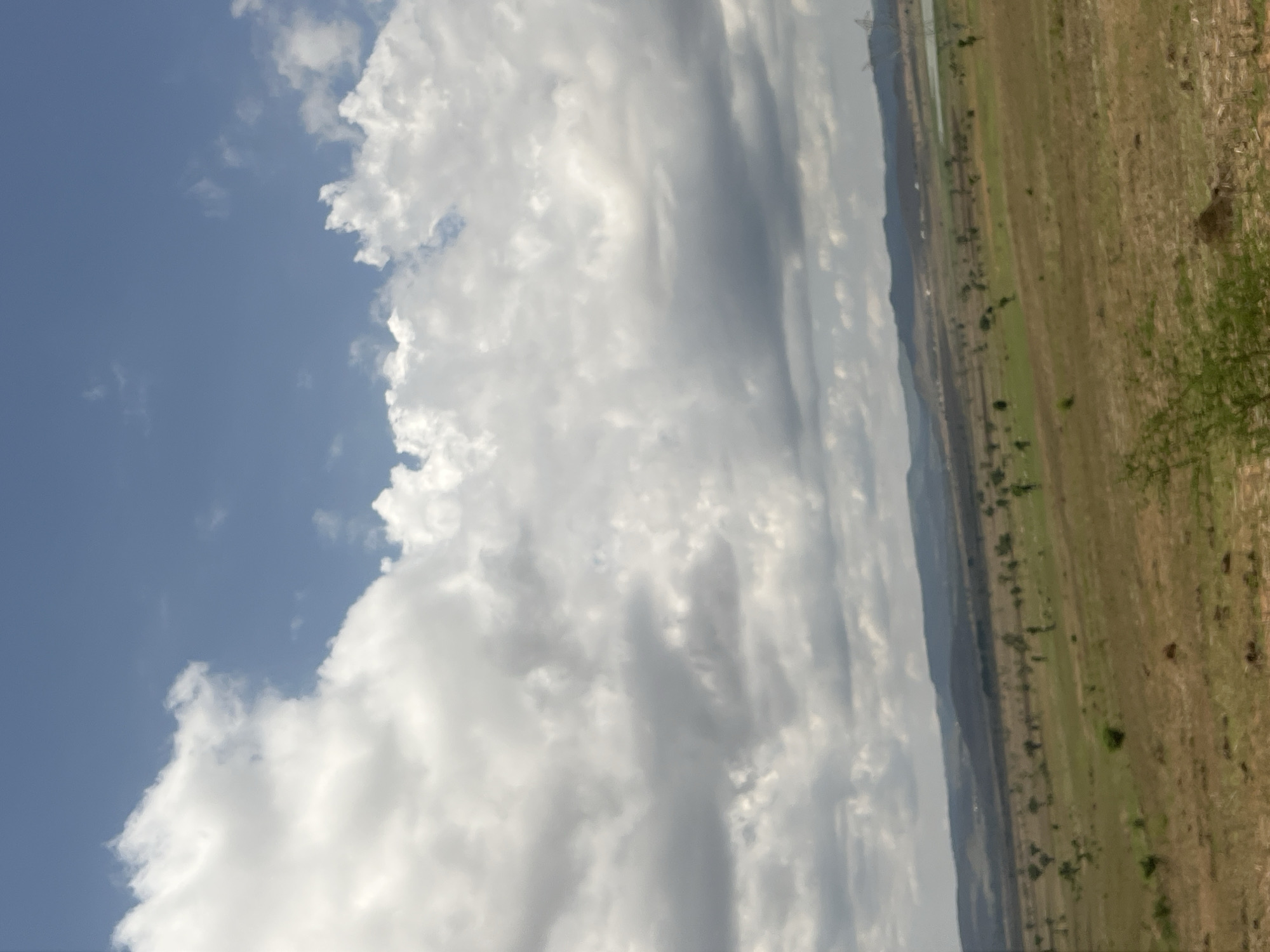 The image displays a wide-angle landscape photograph captured in or near the city of Chum, Nigeria. The upper half of the frame is dominated by a blue sky with extensive cloud cover. Large, white cumulus clouds are prominent on the left, while the clouds transition to a more dispersed and grayish pattern towards the right and center. The lower half of the image presents an expansive, undulating terrain. A distant range of hills and mountains forms the horizon line, displaying varied natural vegetation. The mid-ground and foreground consist of open land, characterized by a mix of dry, brownish grass and patches of greener vegetation, with scattered small trees and shrubs dotting the landscape. In the far mid-ground, to the right, a body of water, likely a small lake or pond, is visible, and further right, a large power line pylon stands against the distant hills. No human or animal subjects are present. The scene conveys a vast natural environment under a partly cloudy sky.